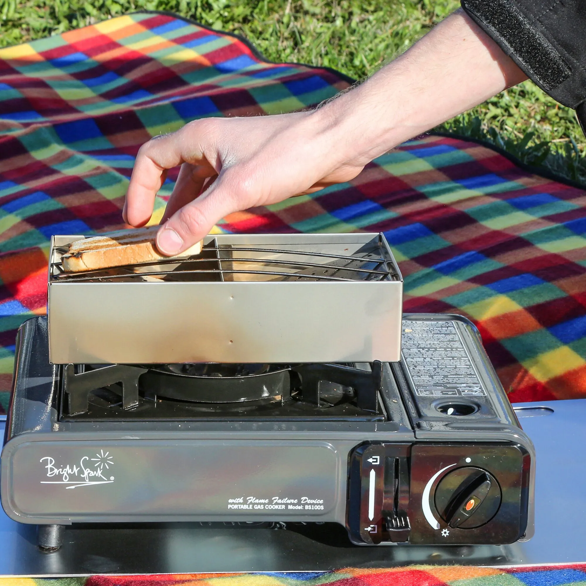 CAMP-A-TOASTER® aus Edelstahl mit Toast auf einem tragbaren Gaskocher im Freien, während eine Hand ihn bedient