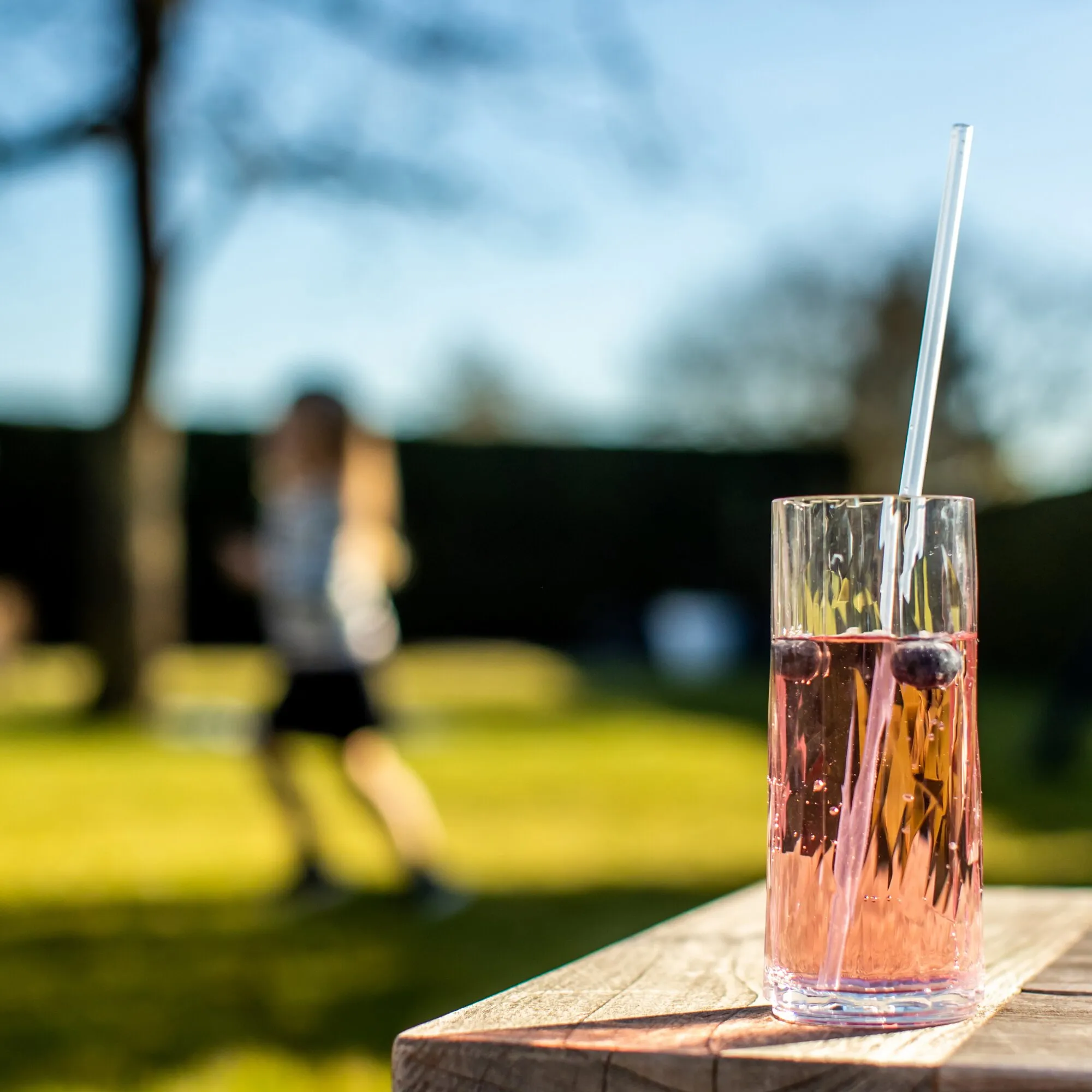 Transparentes Trinkhalm-Set CHEERS in einem Glas mit rosafarbenem Getränk und Beeren auf einem Holztisch im Freien