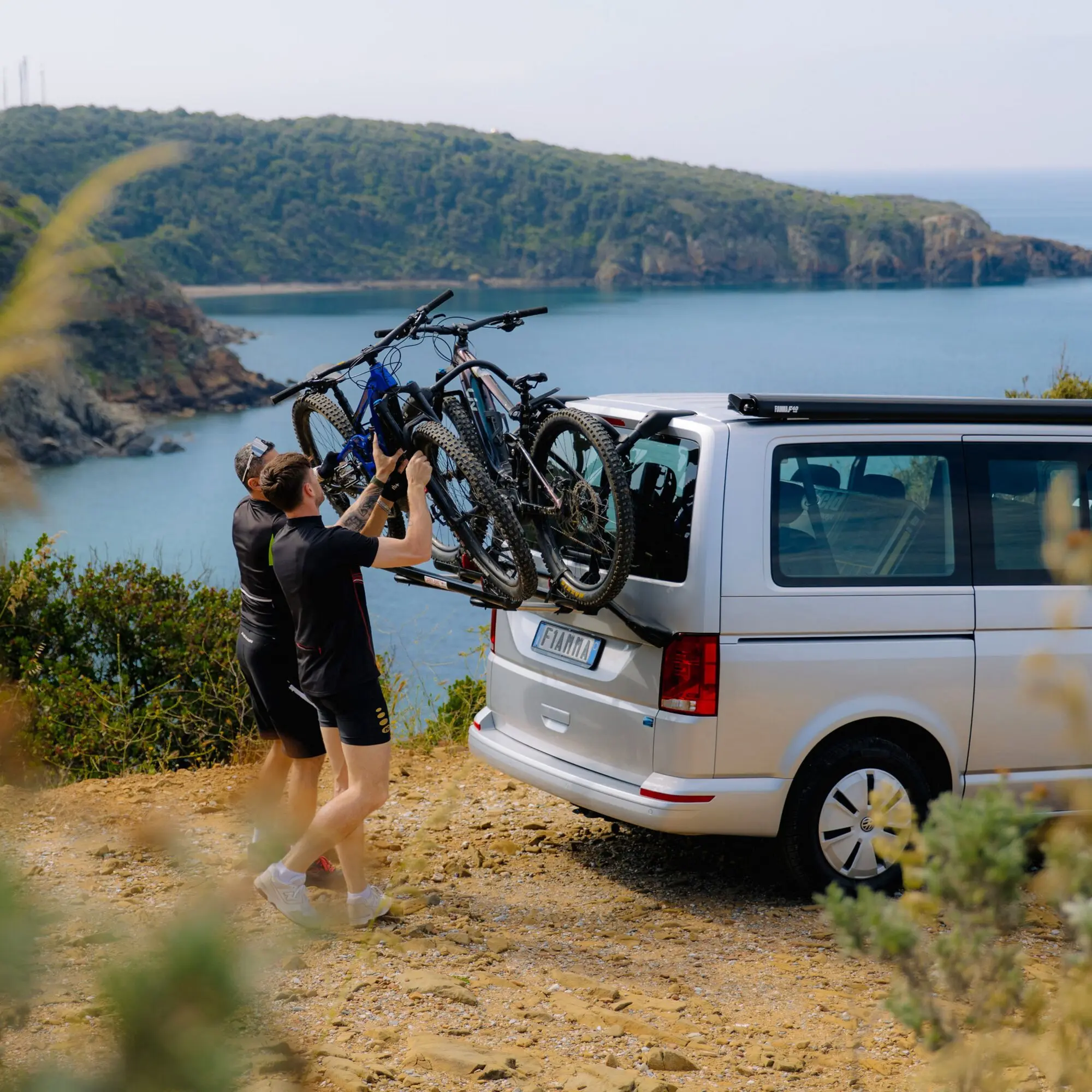 Zwei Mountainbikes werden auf den Heckträger eines Vans gehoben, von zwei Personen unterstützt, vor einer Küstenlandschaft mit Meer und Felsen