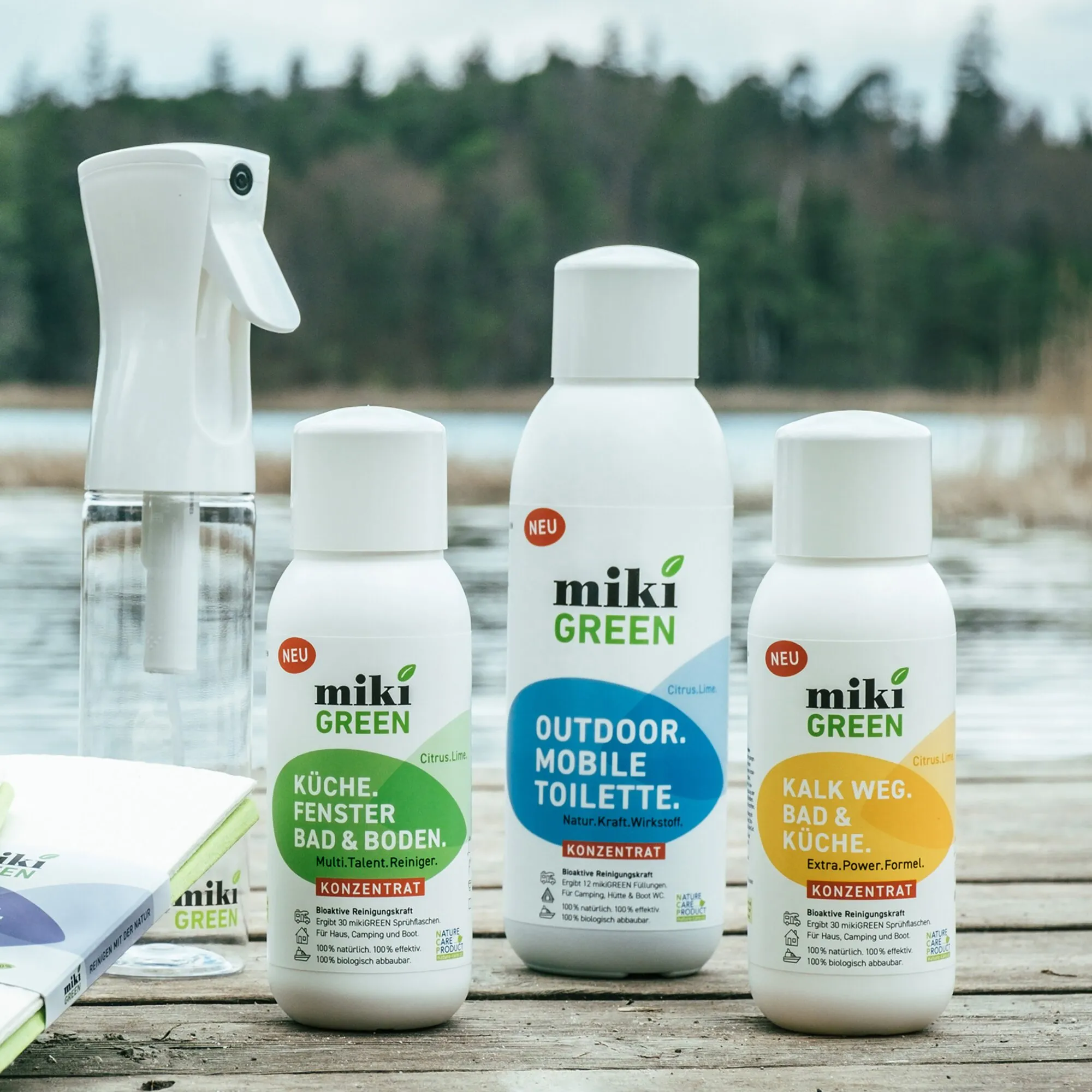Weiße Miki.Misch.Flasche mit Sprühkopf und klarem Behälter neben drei weißen mikiGREEN-Reinigungskonzentraten am See