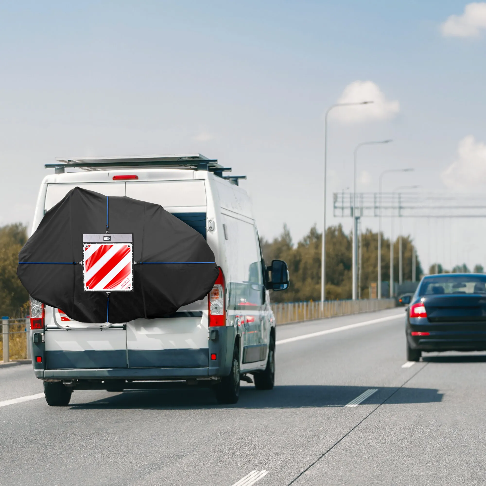 Rückseite eines weißen Transporters mit einer großen schwarzen Fahrradschutzhülle über dem Heck, vermutlich zum Schutz mehrerer Fahrräder, Dachträger
