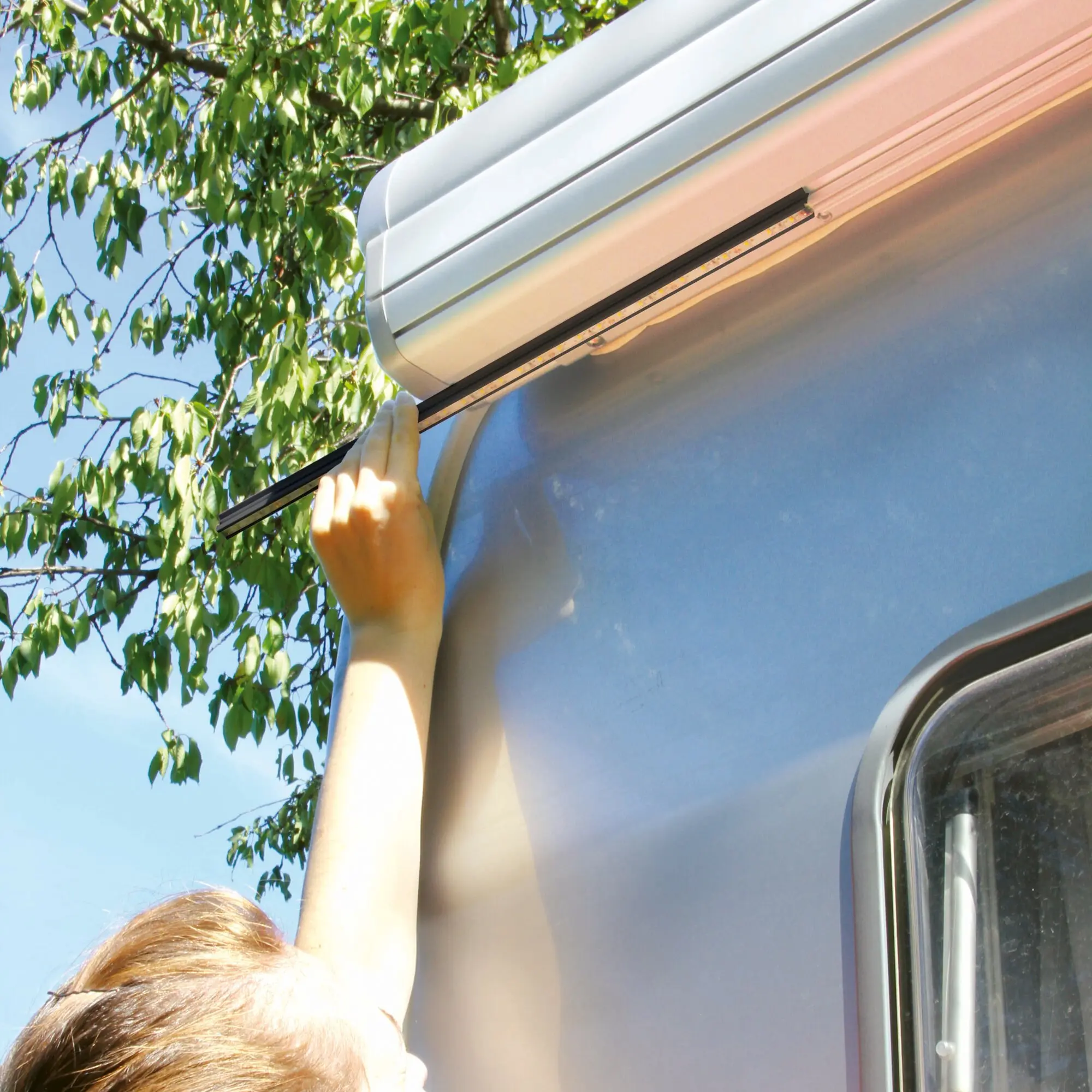 Eine Person montiert eine schwarze Lichtleiste am Awning-Case eines silbernen Wohnmobils, LED-Streifen sichtbar, grüne Blätter im Hintergrund