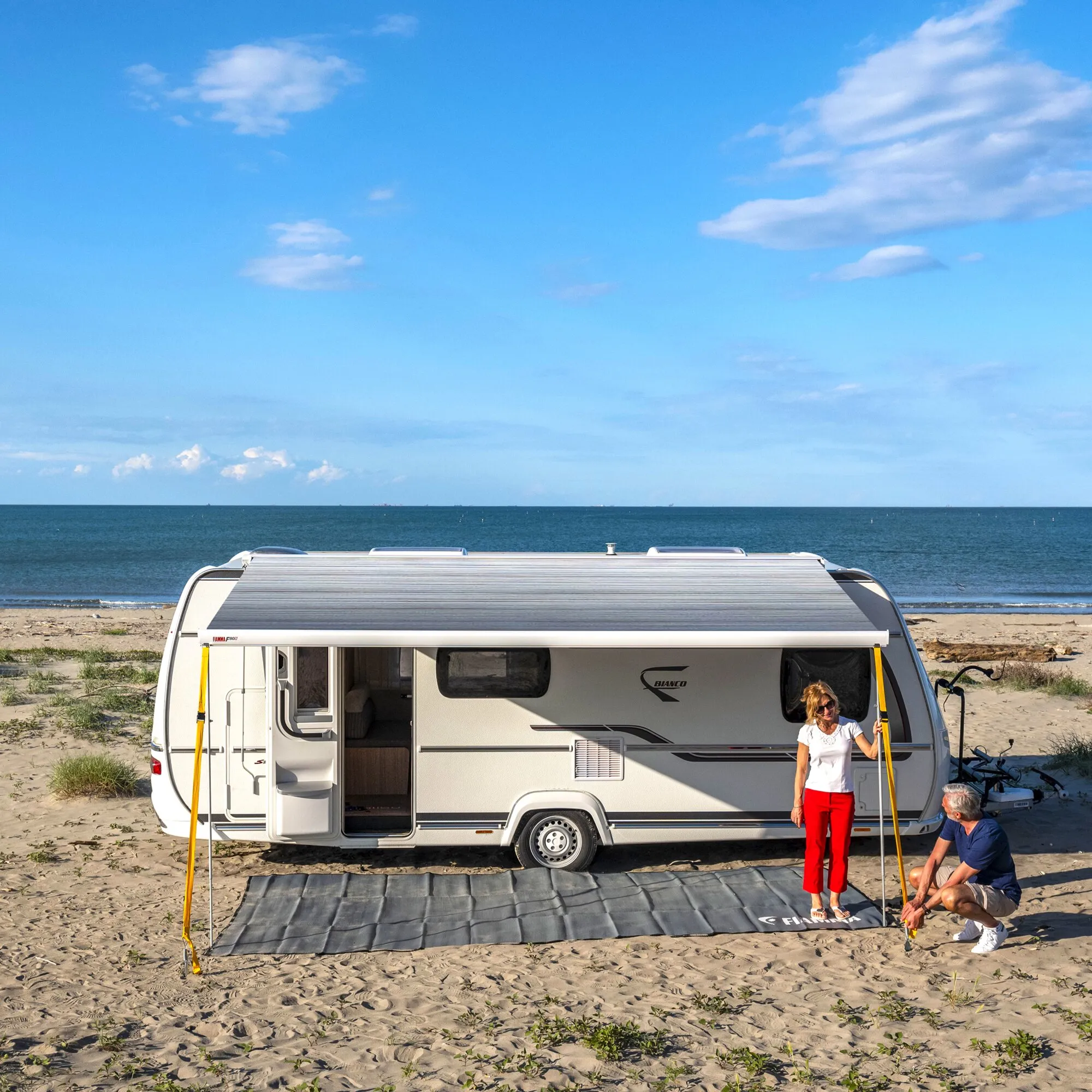 Markise F80 in Polarweiß mit Royal-Grey-Tuch an einem Wohnwagen, am Strand ausgefahren, mit zwei Personen darunter