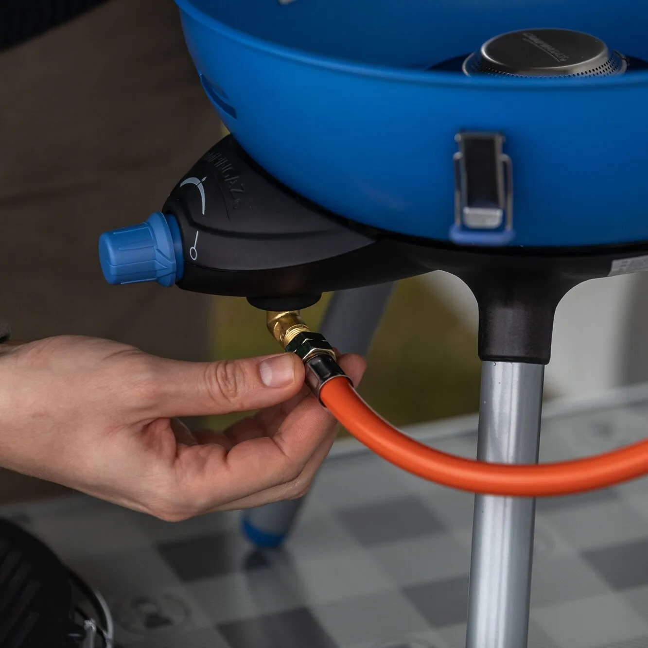 Detailaufnahme des blauen Party Grill 400 Caravan Connect mit orangefarbener Gasleitung, während ein Anschluss per Hand befestigt wird