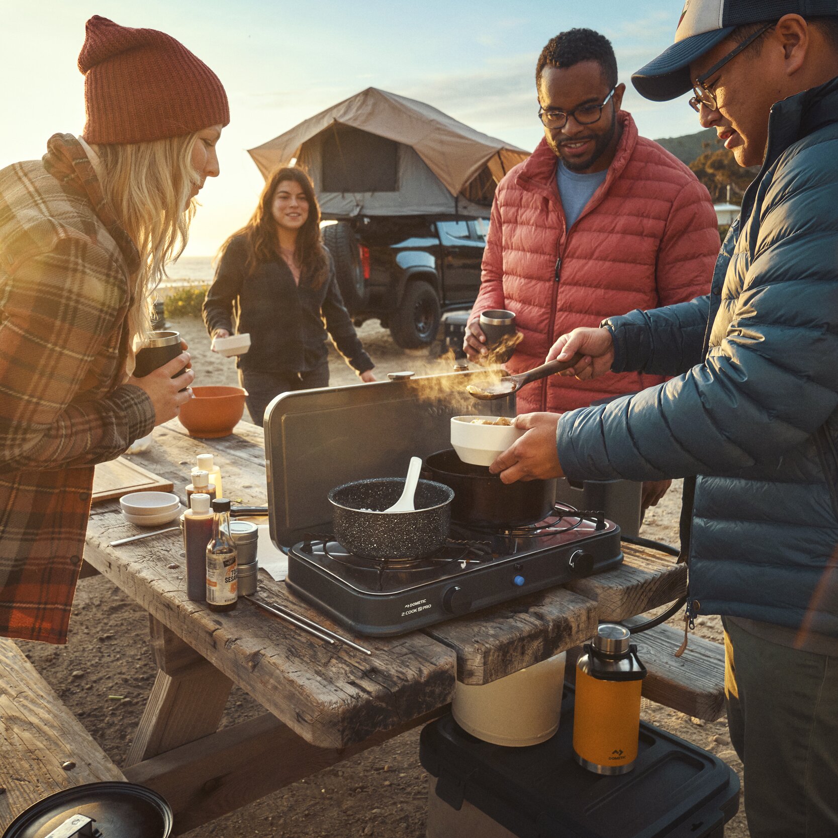 Vier Personen kochen am Campingkocher Van Cook FFD BBQ Plate auf einer Holztafel, neben ihnen gelbe Gasflasche, dampfender Topf und Becher, Zelt im Hintergrund