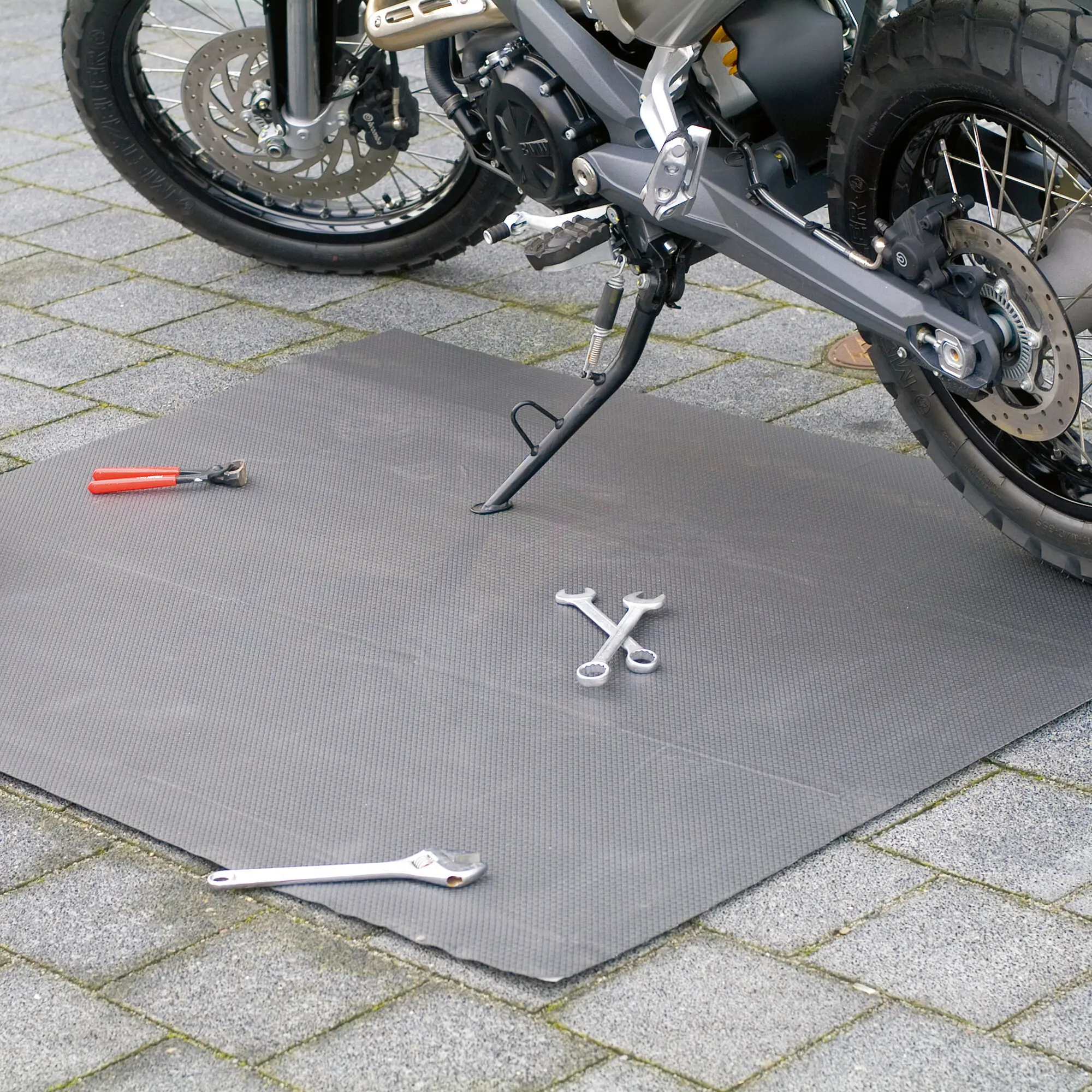 Große dunkelgraue Schmutzfangmatte liegt auf gepflastertem Boden; daneben Schraubenschlüssel und Zange, sichtbar ein Motorrad mit Disc-Bremse