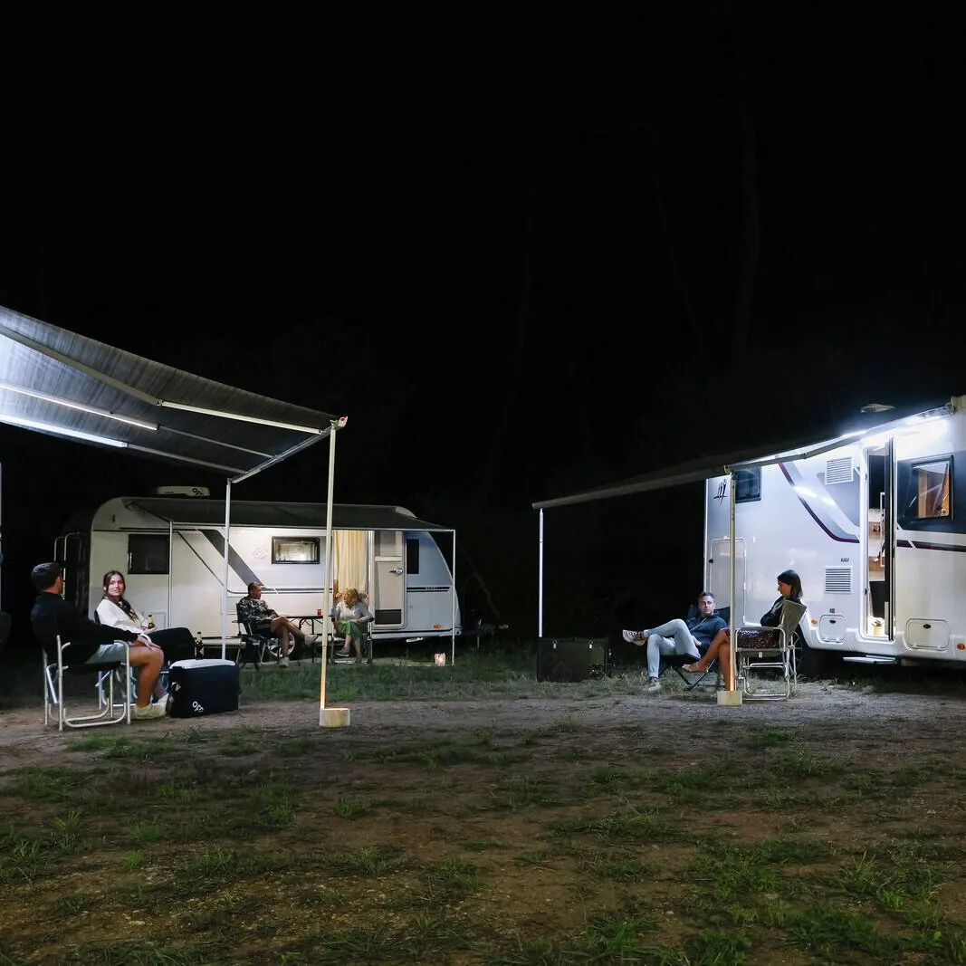 Nächtliche Camping-Szene mit zwei Wohnwagen, ausgefahrenen Markisen und sichtbaren LED-beleuchteten Spannstangen, davor sitzende Camper