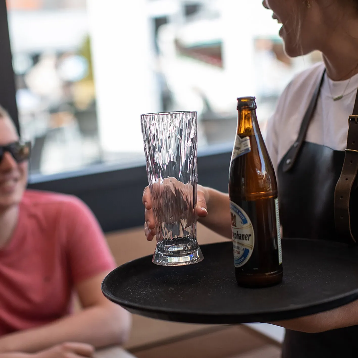 Person hält ein transparentes, strukturiertes Weißbierglas Superglas CLUB neben einer geöffneten Bierflasche auf einem Serviertablett
