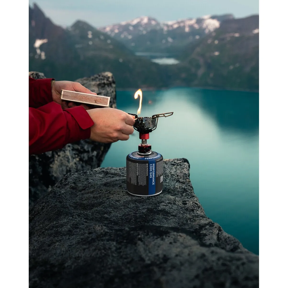 Schwarze 240-g-Gaskartusche C 300 mit Campingkocher und Flamme auf einem Felsen vor einem Bergsee