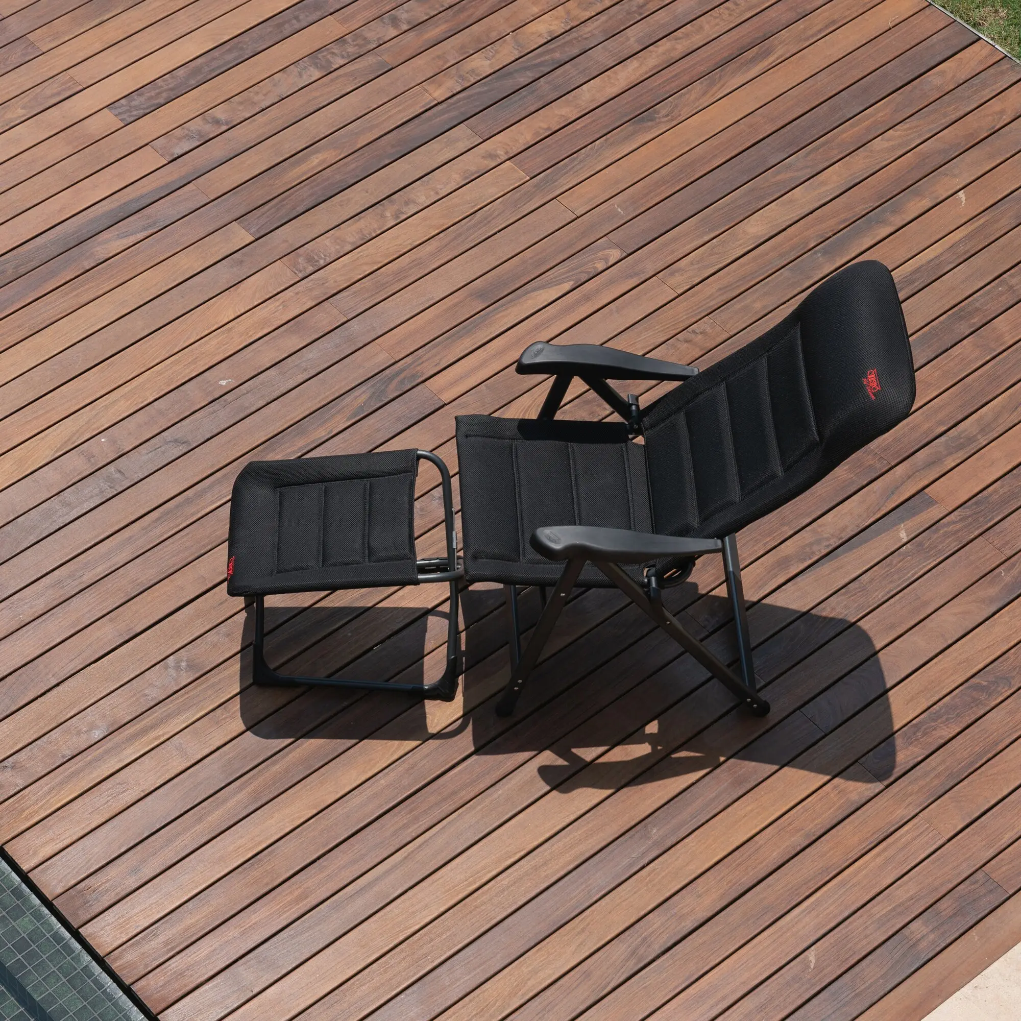 Schwarze Campingstühle mit Gewebe-Sitze und Metallrahmen stehen auf braunem Holzdeck; ein rotes Logo ist am Rückenpolster sichtbar