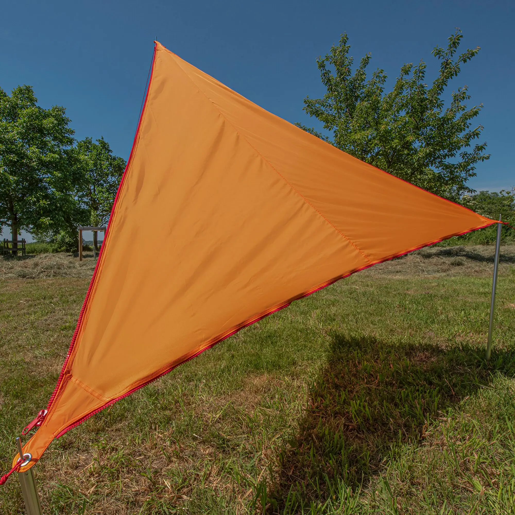 Orangfarbenes Sonnensegel Zip Canvas mit roter Nahtkante, dreieckige Form, an Metallpfosten befestigt, Hintergrund aus blauem Himmel und grünen Bäumen