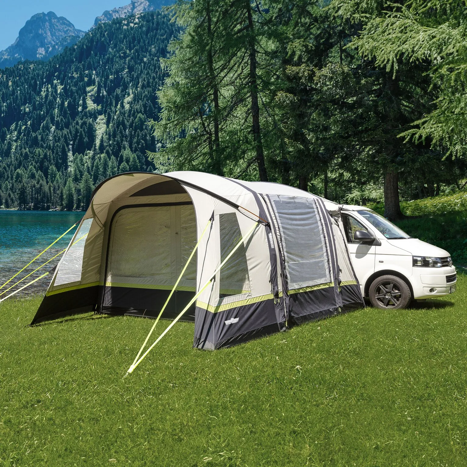 Busvorzelt Advantourer in Grau und Oliv, an einem weißen Kleinbus auf einer Wiese an einem Bergsee aufgebaut