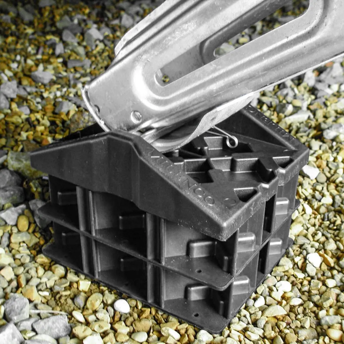 Schwarzer Kunststoffblock mit Verstrebungen, darauf liegt eine silberne Metallstützplatte mit Langloch und Befestigungslöchern; Kieselsteine