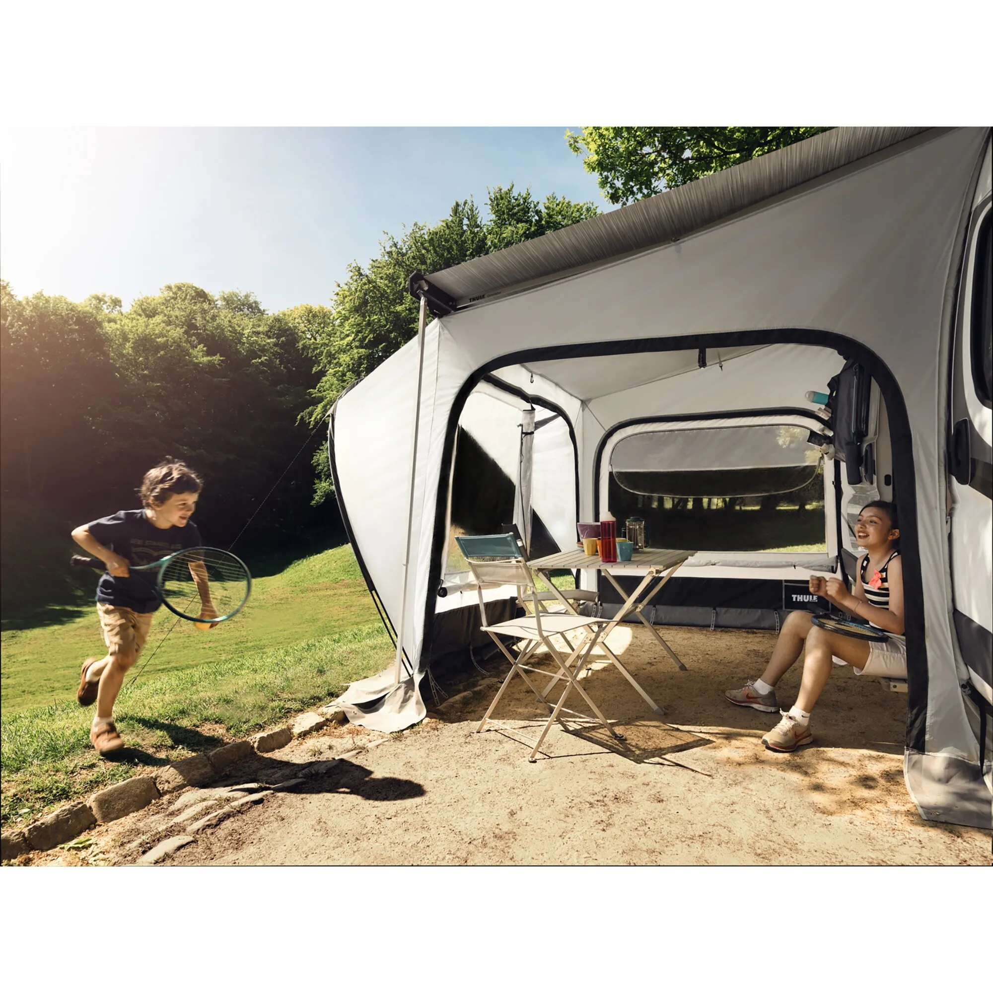 Weißes Markisenzelt Thule QuickFit Ducato an einem Campingbus, mit Tisch und Stühlen; ein Kind spielt davor, eine Frau sitzt im Zelt