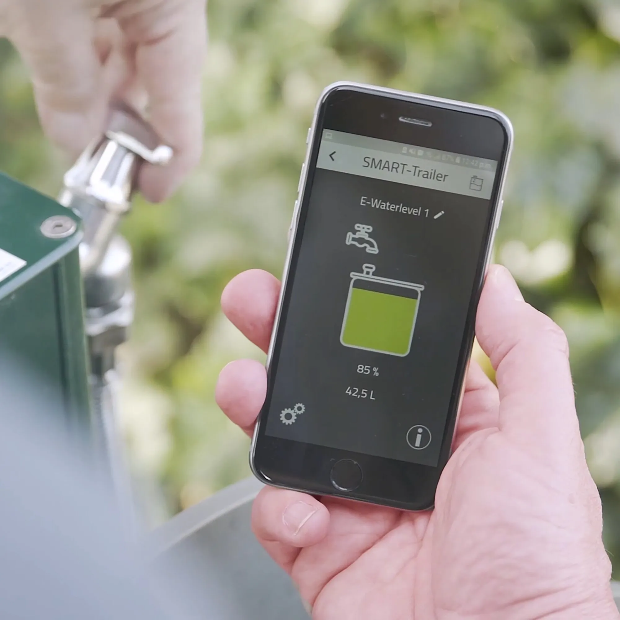 Hand hält ein Smartphone mit E-Waterlevel-App, die 85 Prozent und 42,5 Liter Wasserstand neben einem Wassertank anzeigt
