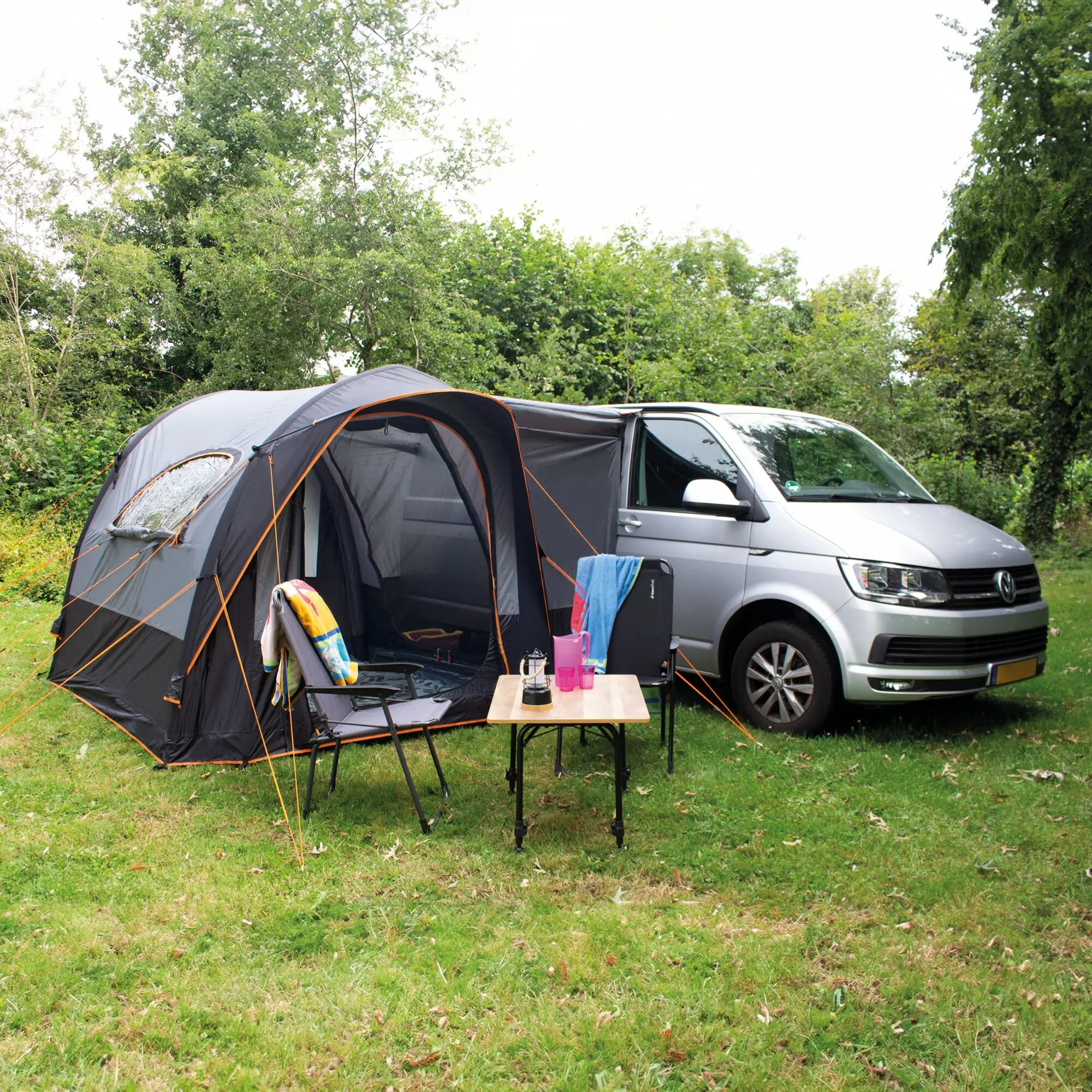 Campingsetup am Van mit grauem Roadster Air Busvorzelt, orange Abspannleinen; Tisch mit Gläsern vor grünem Gras und Bäumen