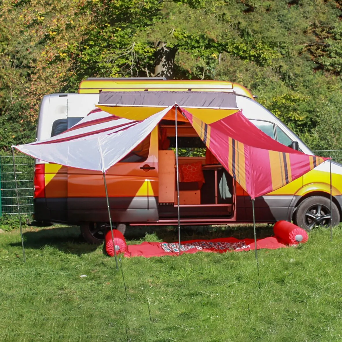 Campingbus mit ausgefahrenem rot-gelb gestreiftem Vorzelt auf einer Wiese, Nutzung des Universal Fahrzeugadapters XL