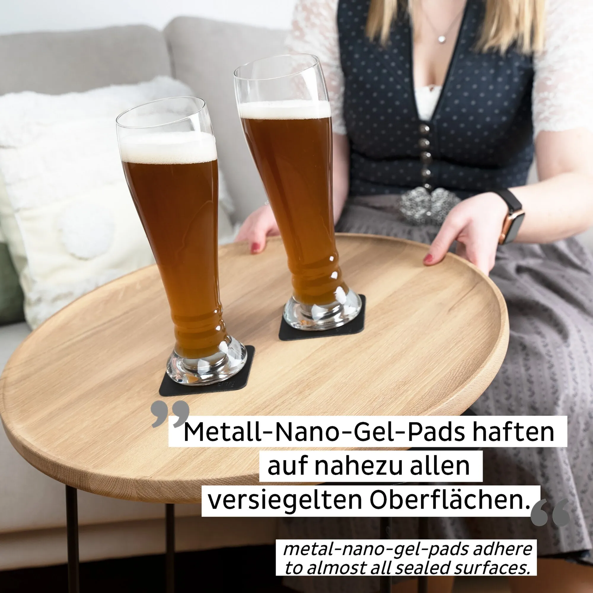 Zwei transparente Magnet-Kristallgläser mit braunem Weißbier und Schaum auf schwarzen Nano-Gel-Pads auf einem Holztisch, daneben eine Person