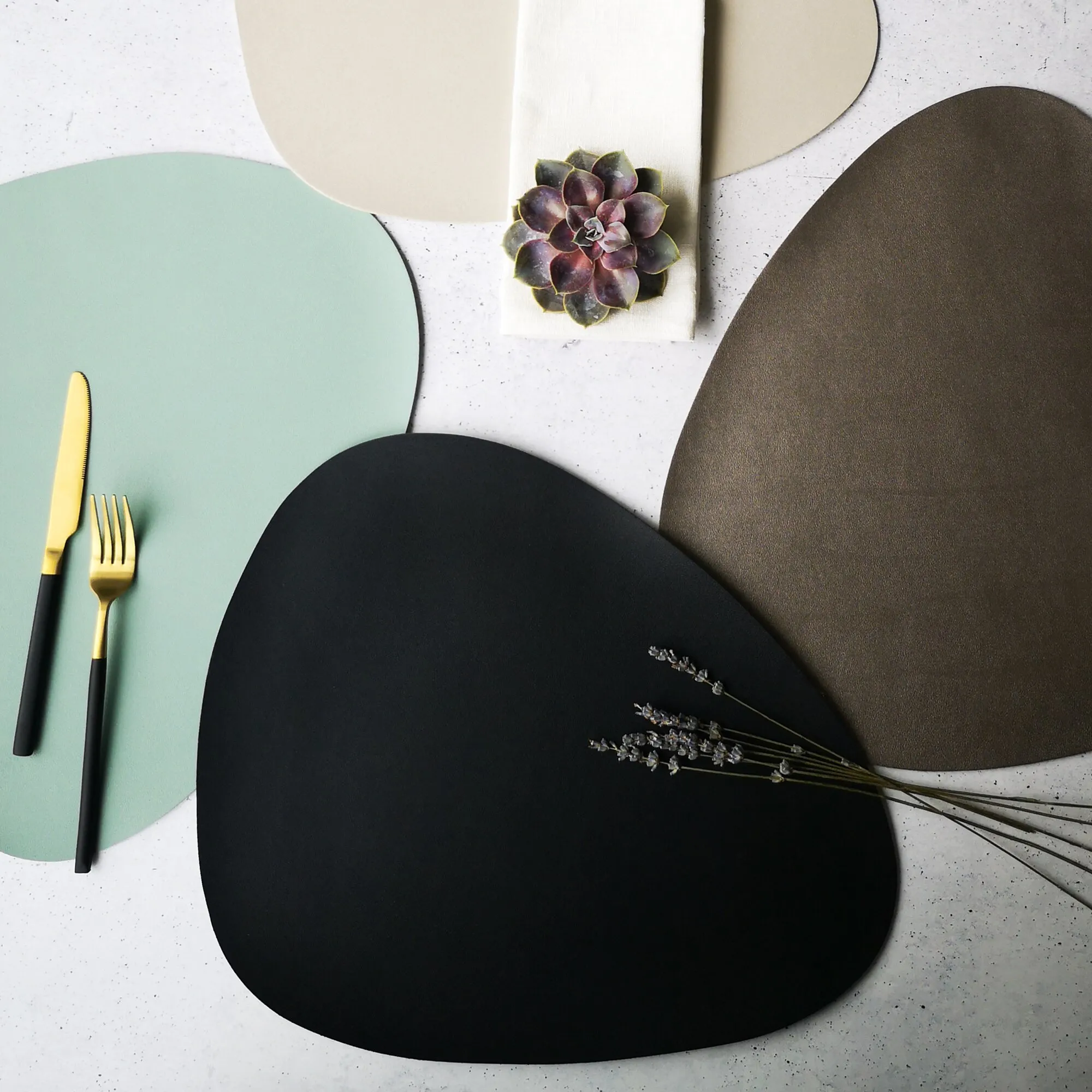 Schwarzes, organisch geformtes Platzset auf einem gedeckten Tisch mit Tellern, gold-schwarzem Besteck und Lavendelzweigen