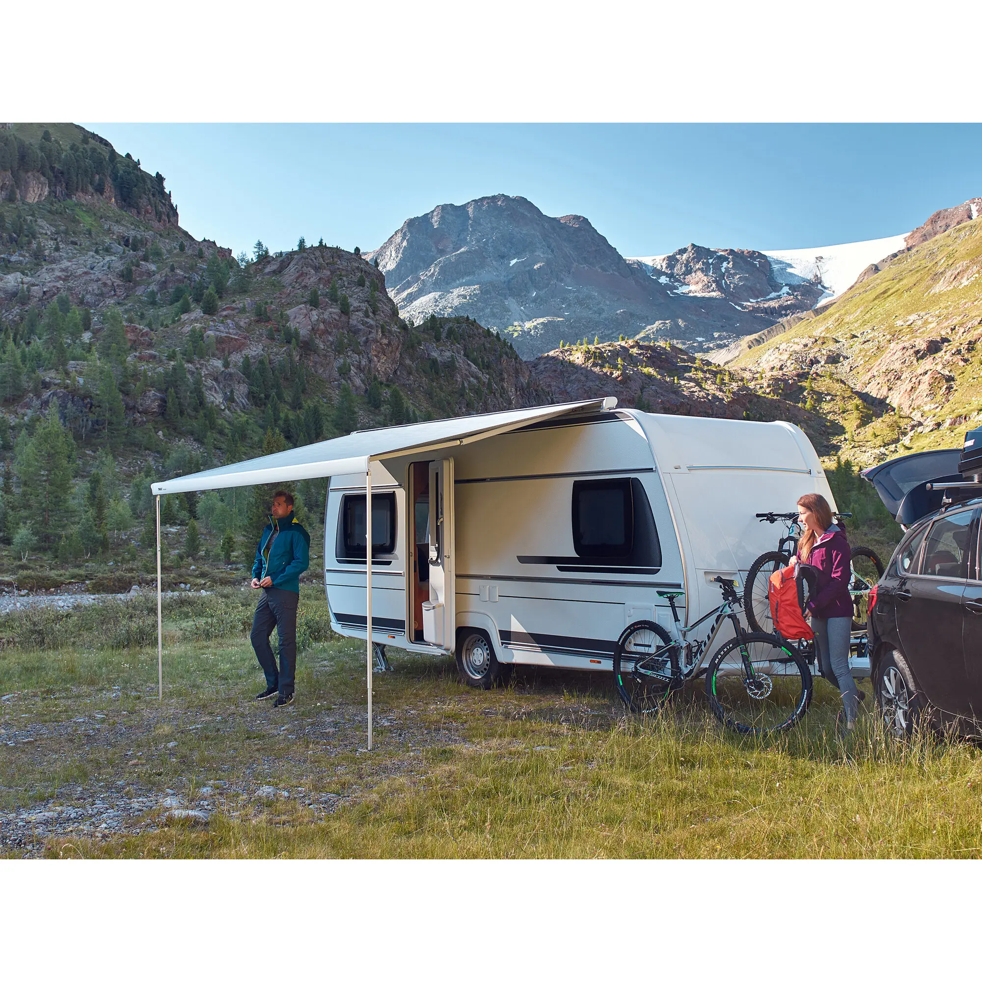 Ausgefahrene anthrazitfarbene Markise Thule Omnistor 6300 am Wohnmobil, motorisiert, 4 m, davor zwei Personen mit Fahrrädern vor Berglandschaft