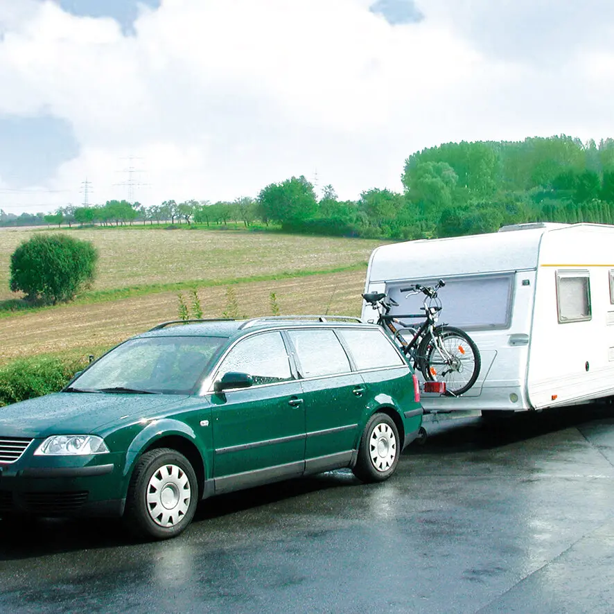 Grüner Kombi zieht einen weißen Wohnwagen; hinten am Wohnwagen ist eine befestigte Fahrradträgeraufnahme sichtbar, auf der zwei Fahrräder montiert sind