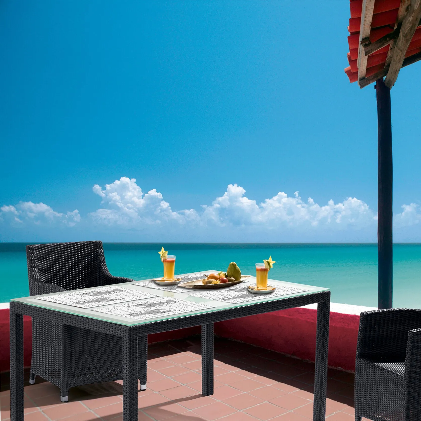 Mehrere weiße Tischsets Miami mit grauem Schriftmotiv liegen auf einem schwarzen Gartentisch am Meer, mit Getränken und Obst gedeckt
