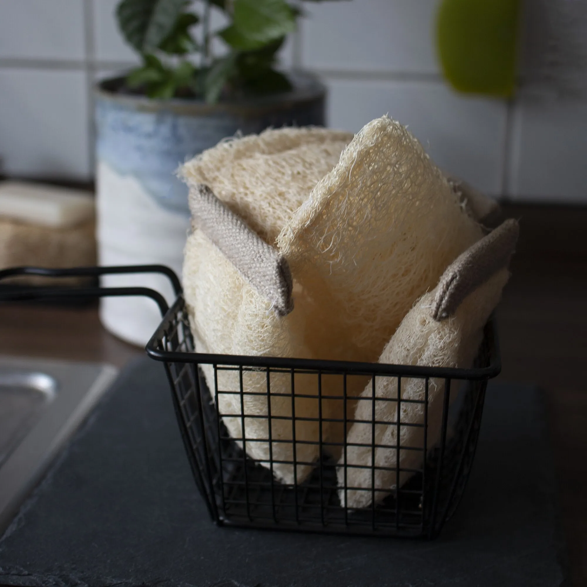 Mehrere cremefarbene Luffa-Schwämme liegen in einem schwarzen Drahtkorb auf einer Küchenarbeitsplatte