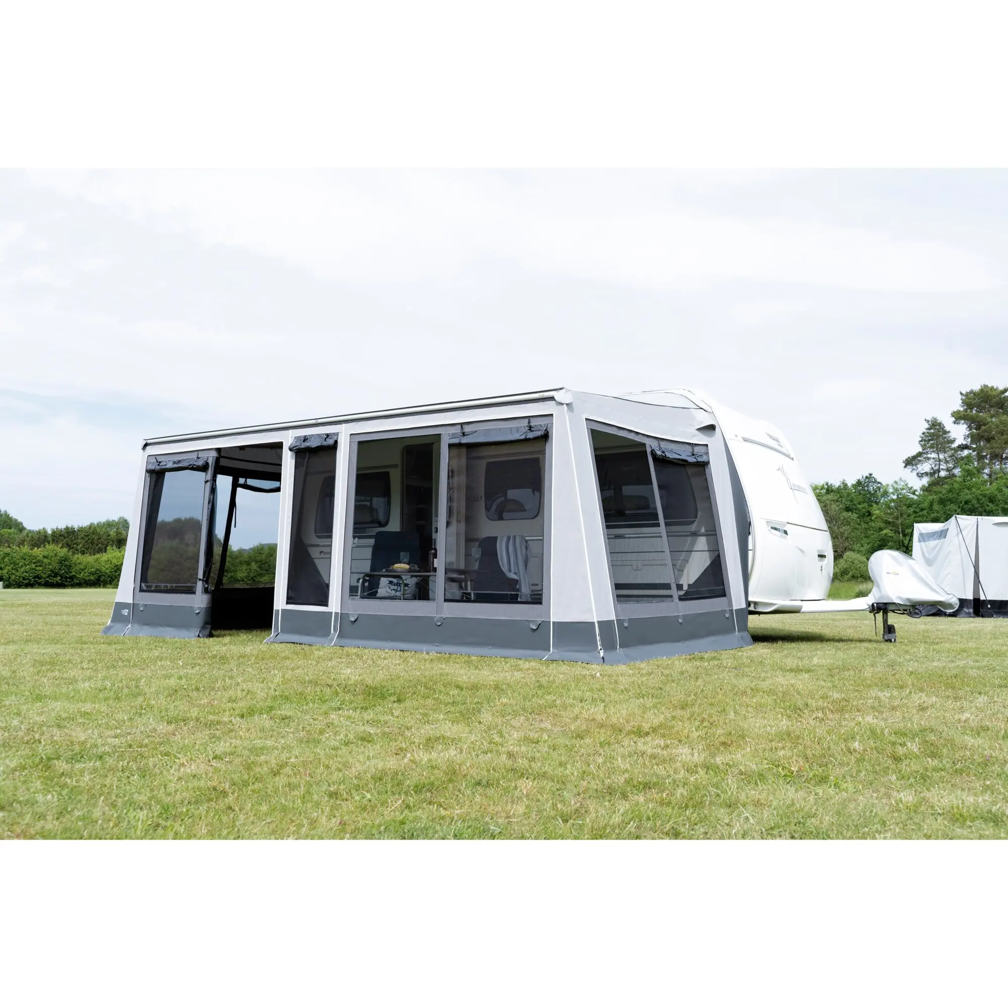 Markisenzelt Rolli Plus Lounge in hellgrau, an einem Caravan befestigt, große seitliche Fensterfronten und offener Eingang, auf grünem Rasen