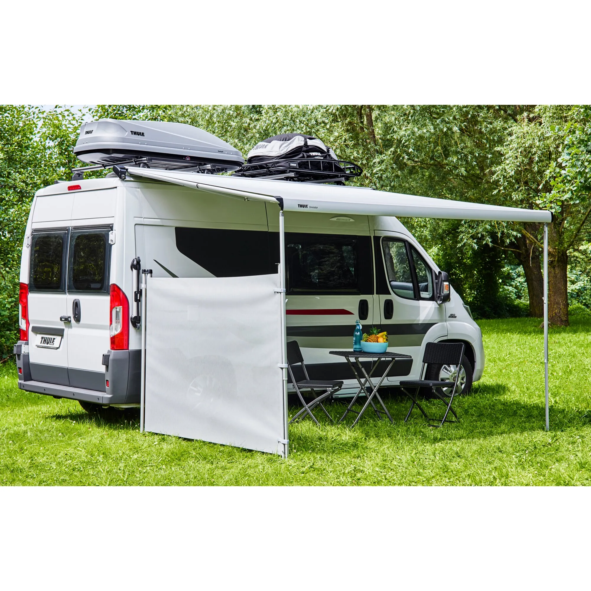 Thule Windscreen als weiße Seitenwand an einer Markise eines weißen Wohnmobils im Grünen, mit Dachbox und Campingmöbeln
