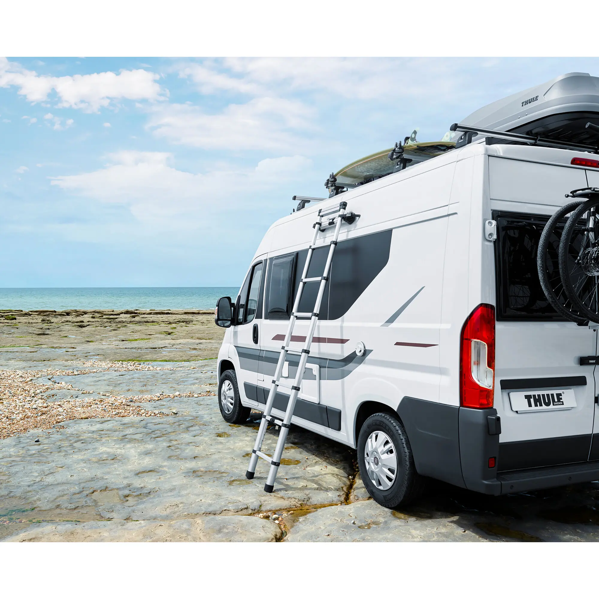 Weißer Camper-Van mit Dachbox von Thule, seitlich montierte Ausziehleiter, Surfbrett auf dem Dachträger, Küstenlandschaft im Hintergrund