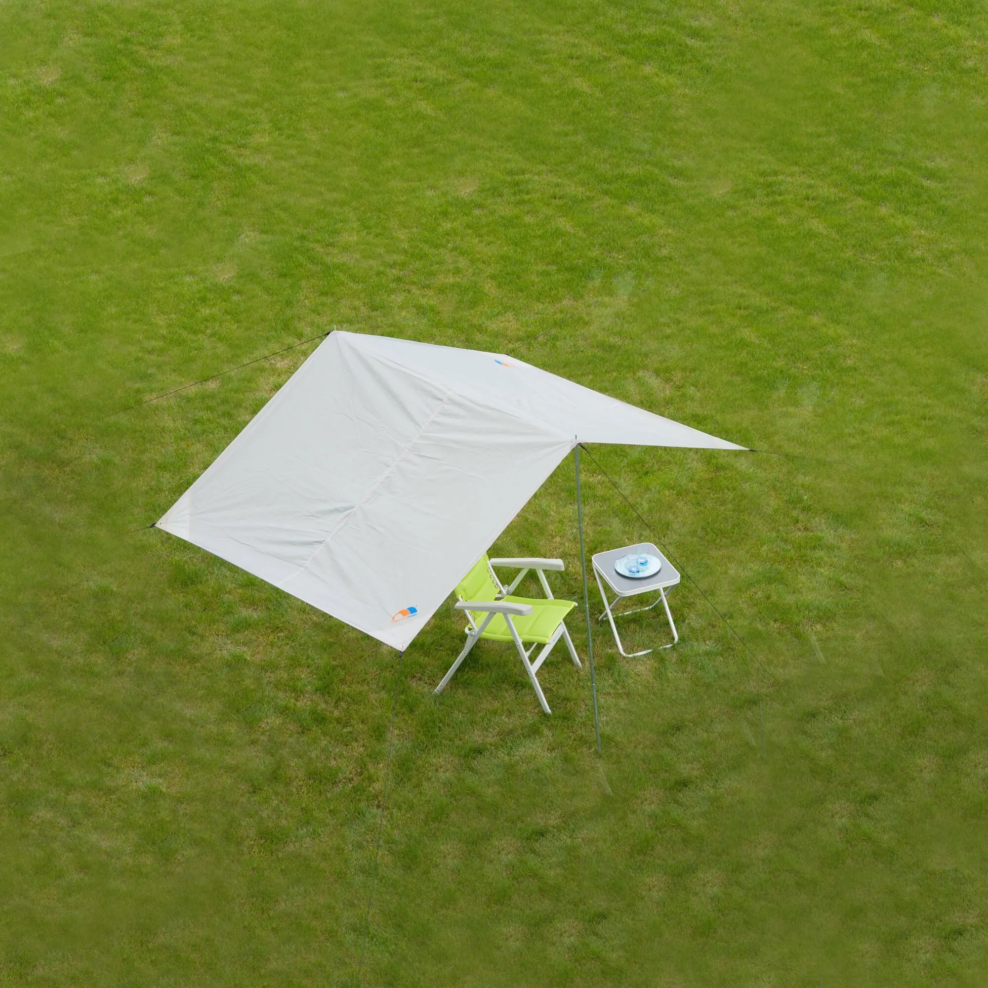 Weißes dreieckiges Sonnensegel Basic auf einer Wiese, mit Abspannleinen und Mast über grünen Campingstühlen und einem Beistelltisch gespannen