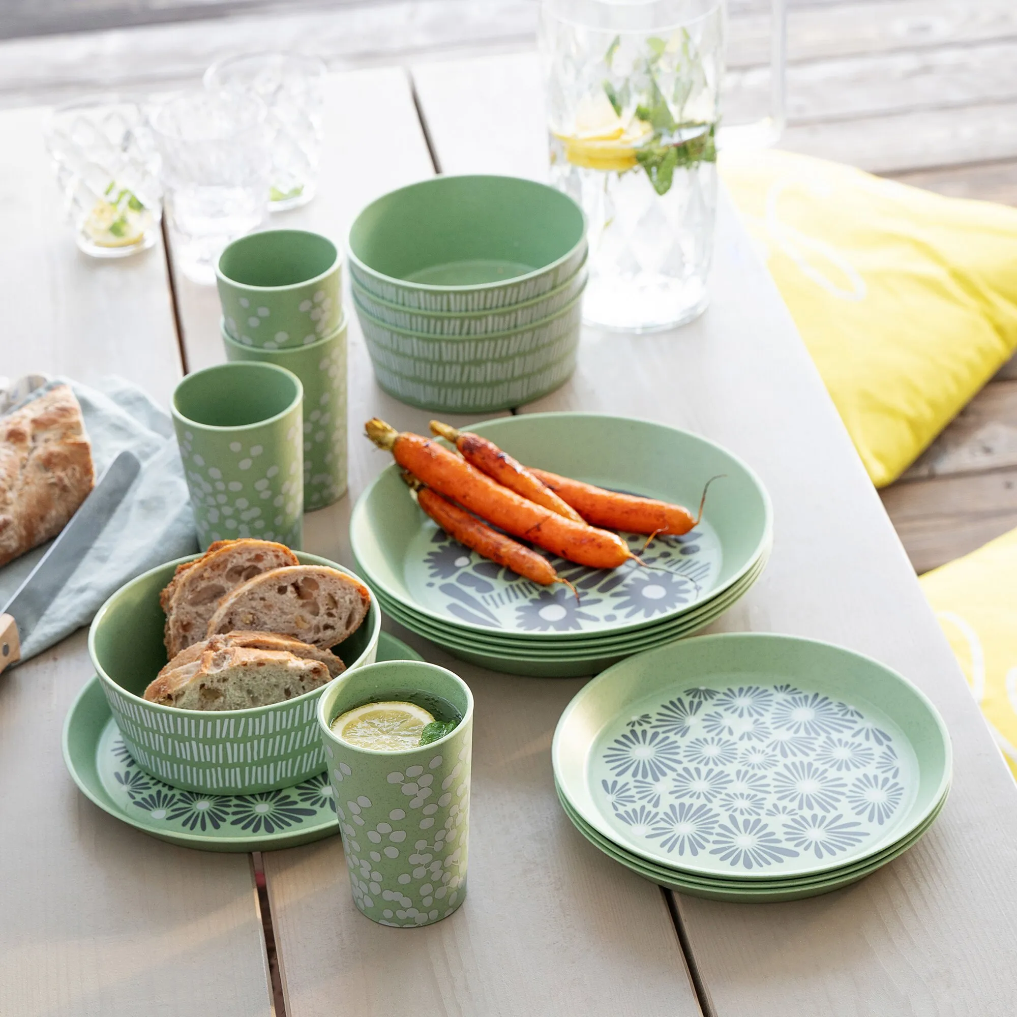 Grünes 16-teiliges Geschirrset mit Blumenmuster: Teller, Schalen und Becher auf Holztisch, daneben Gläser mit Zitronen, Minze und Brot