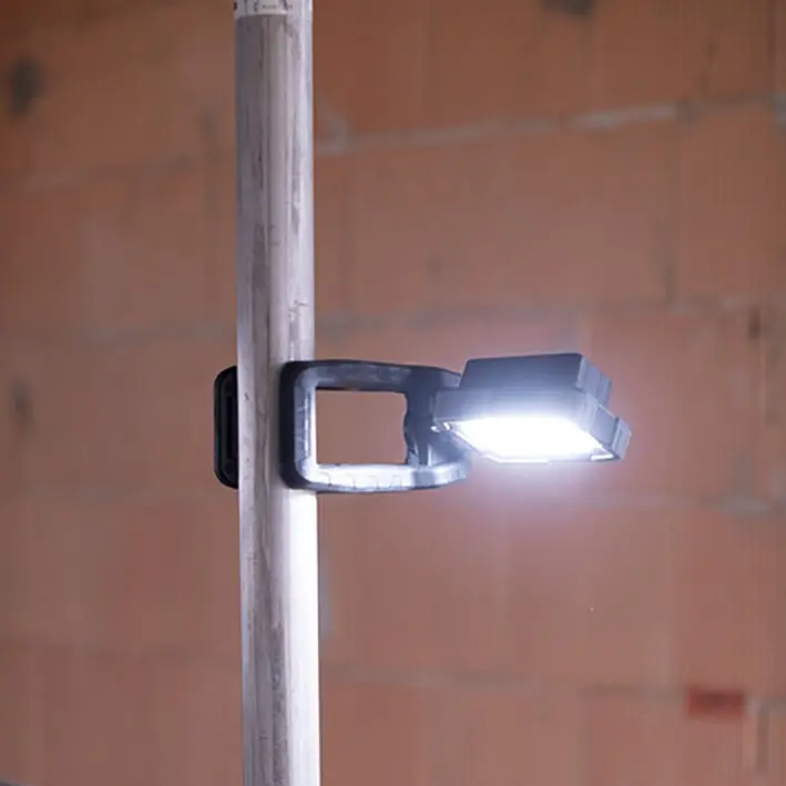 Klemmbarer mobiler LED-Arbeitsstrahler mit rechteckiger Leuchtfläche, an einer Stange befestigt, leuchtet hell, vor brauner Ziegelwand