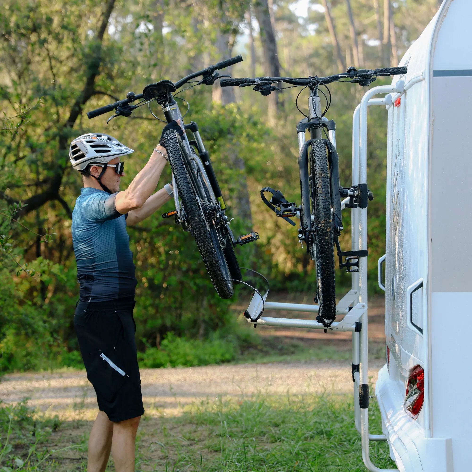 Mann befestigt zwei Mountainbikes am weißen Carry-Bike Universal Fahrradträger eines Wohnmobils im Wald
