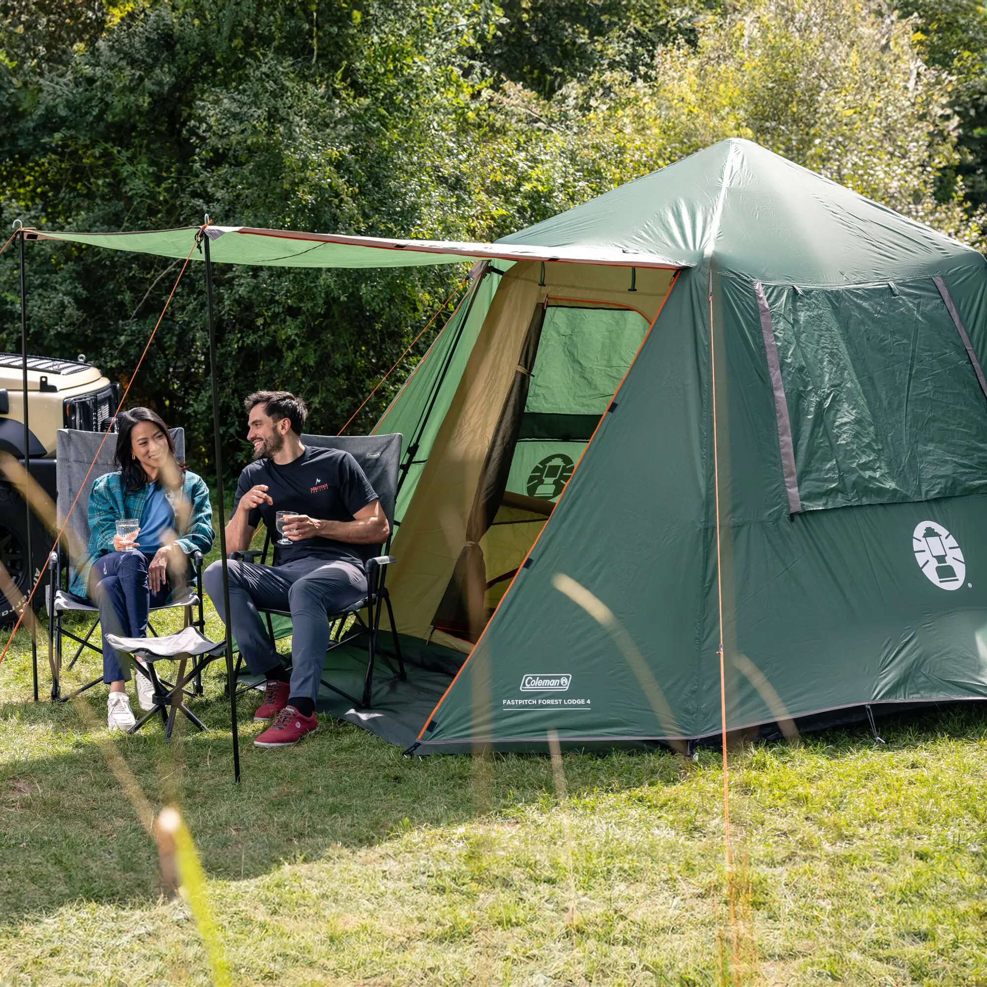 Grünes Kuppelzelt mit offenem Eingang, grüne Markise und orange Abspannleinen, zwei Personen sitzen in Klappstühlen davor, Wald im Hintergrund