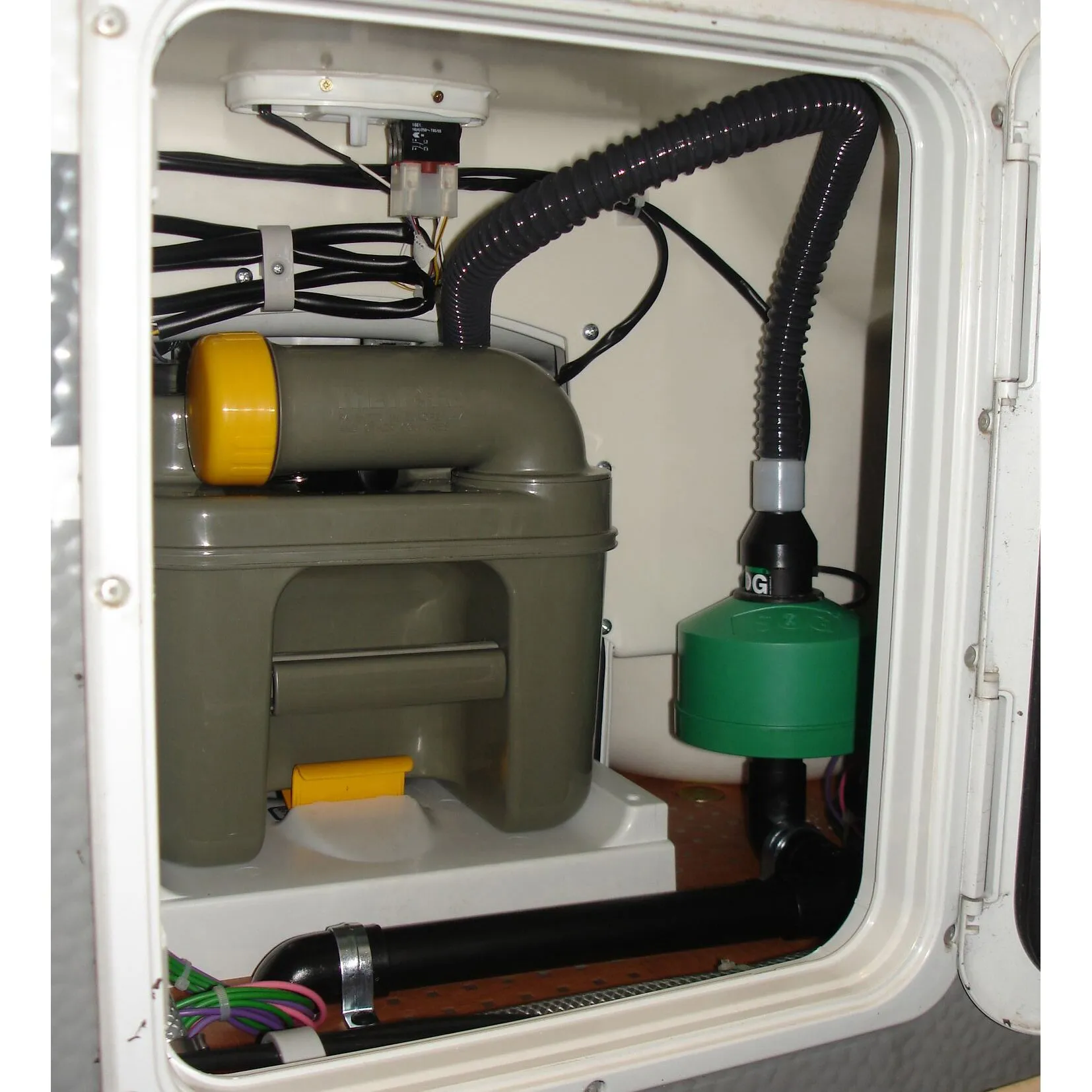 Innenansicht einer weißen C200-WC-Tür mit elektrischer Entlüftung, schwarzem Abluftschlauch, Kabeln und angeschlossenem Fäkalientank