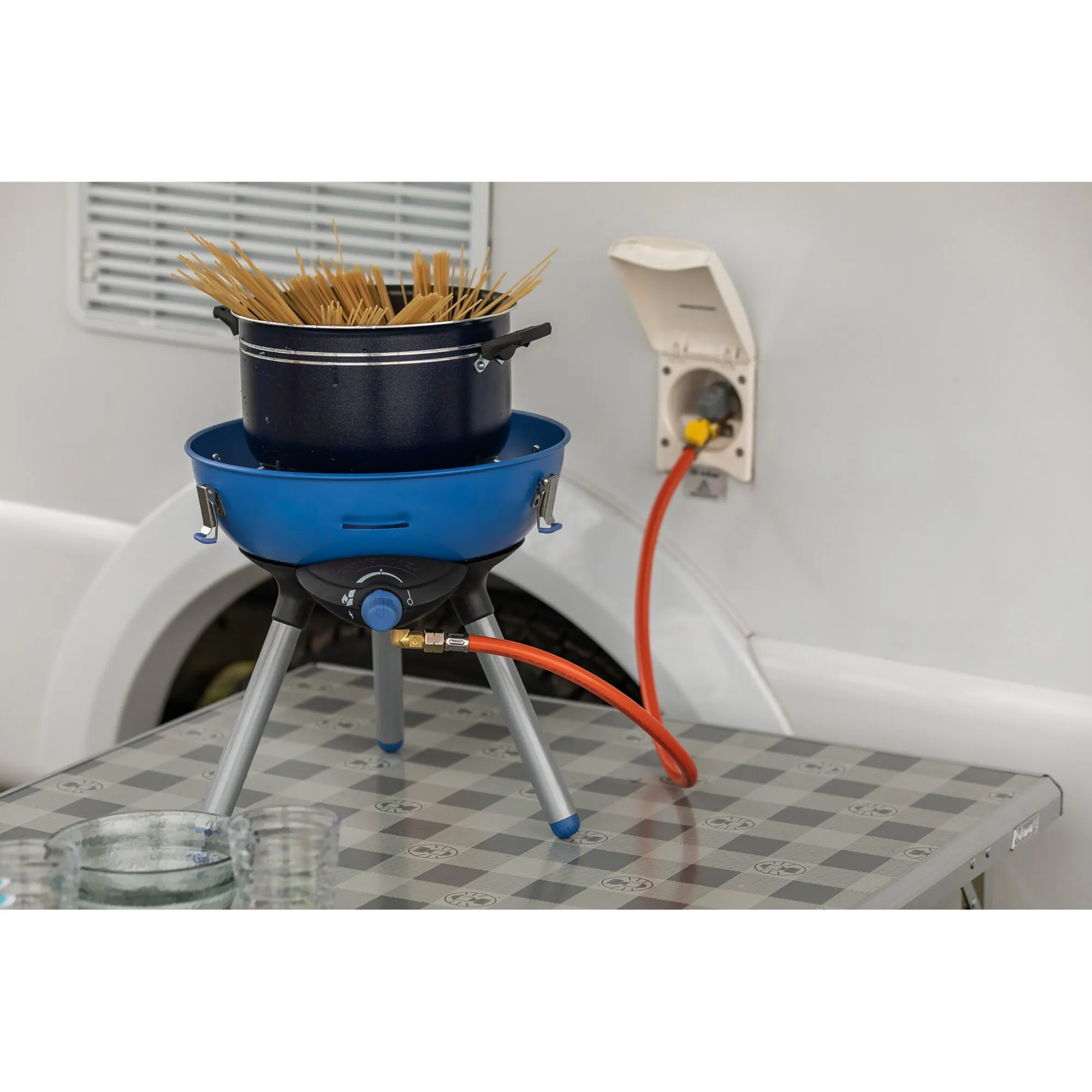 Blauer Party Grill 400 Caravan Connect auf einem Campingtisch, per Gasschlauch am Wohnwagen angeschlossen, mit Topf Spaghetti darauf