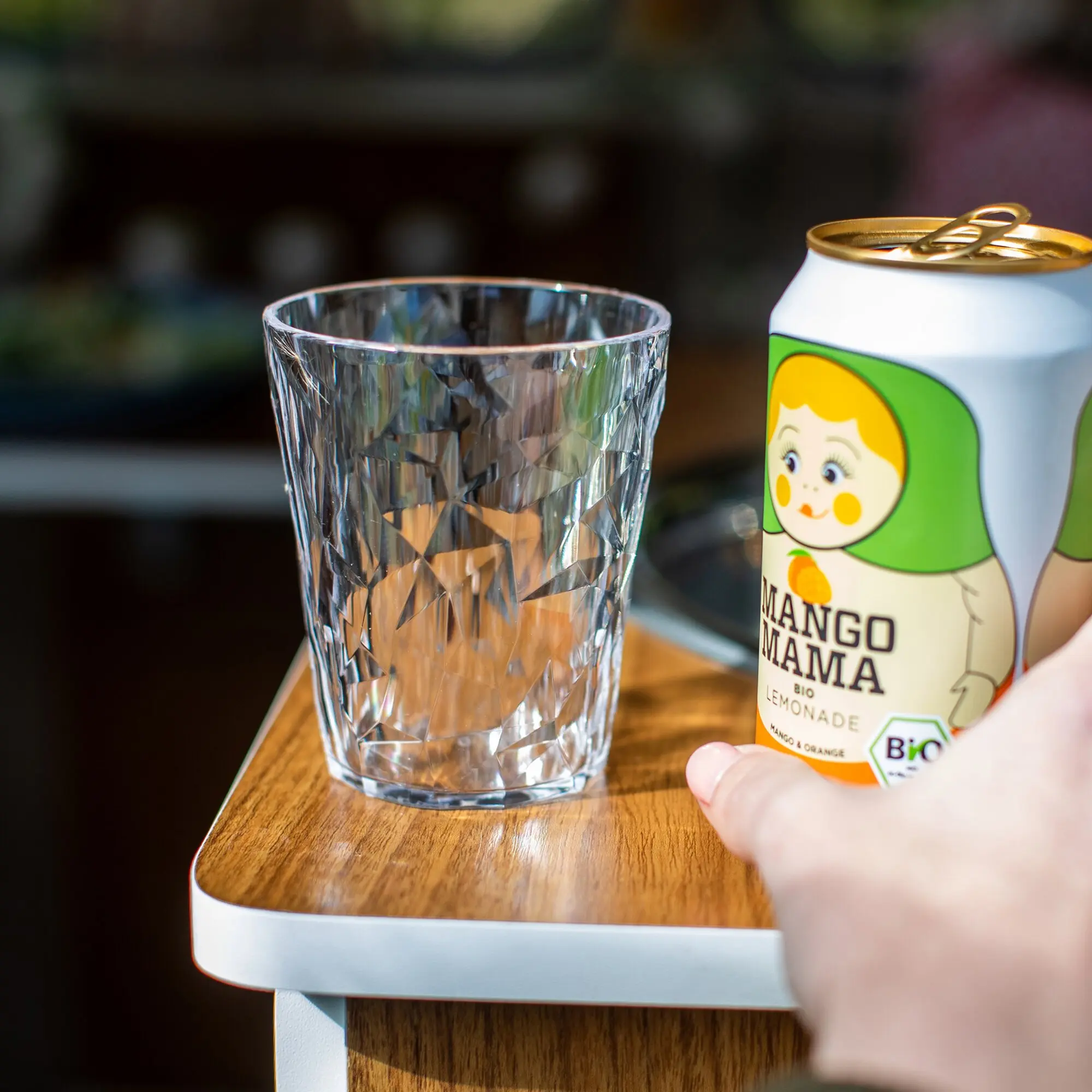 Nahaufnahme eines facettierten Kristallglases auf einem Holztisch, eine Hand hält eine Mango Mama Limonade-Dose mit Cartoon-Gesicht, unscharfer Hintergrund