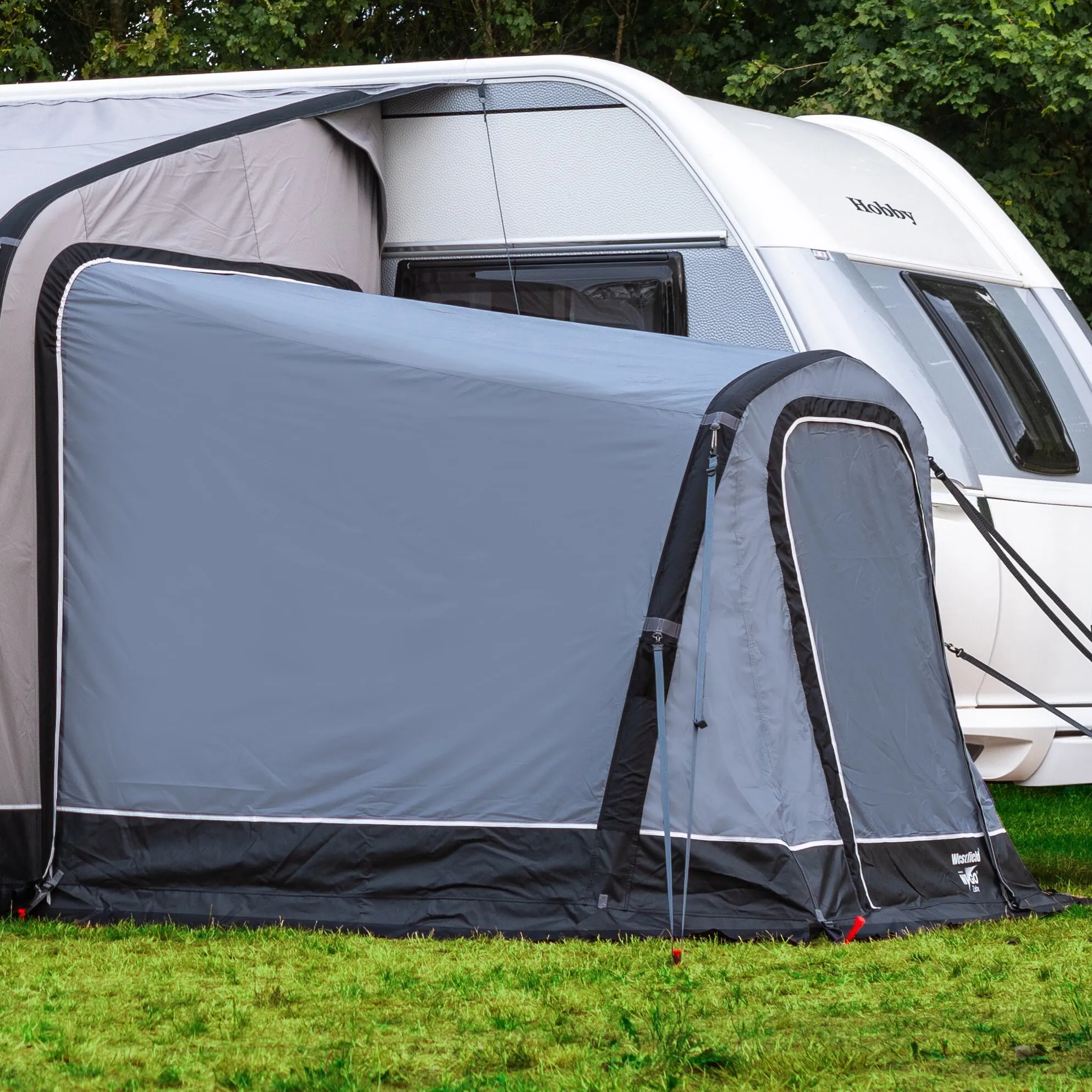 Grauer Anbau Universal für Vorzelte, seitlich an einem weißen Hobby-Wohnwagen aufgebaut, mit Eingang und Abspannleinen auf einer Wiese