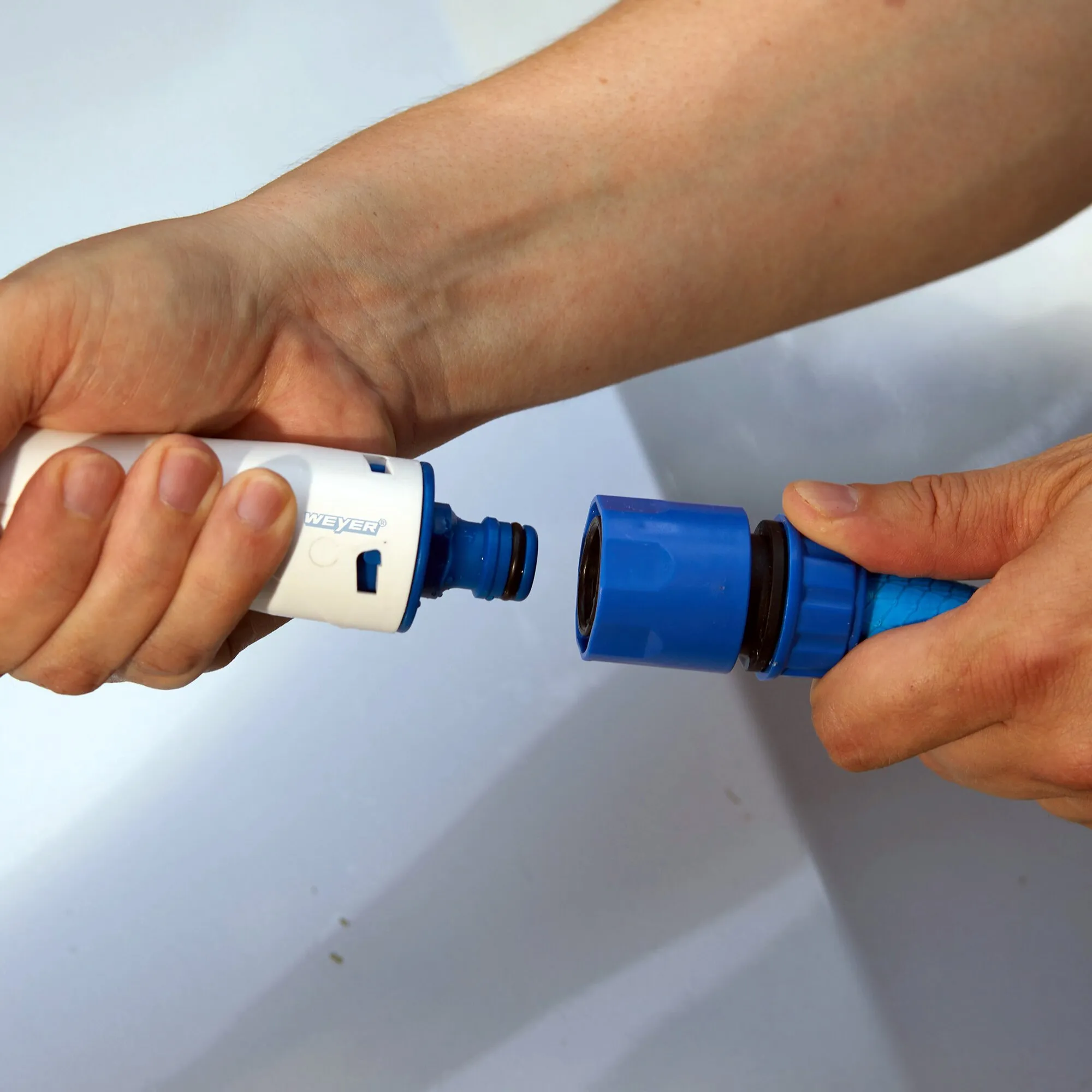 Hände verbinden den weißen Griff einer ovalen Waschbürste mit blauem Anschluss mit einer blauen Schlauchkupplung; der Bürstenkopf ist nicht sichtbar