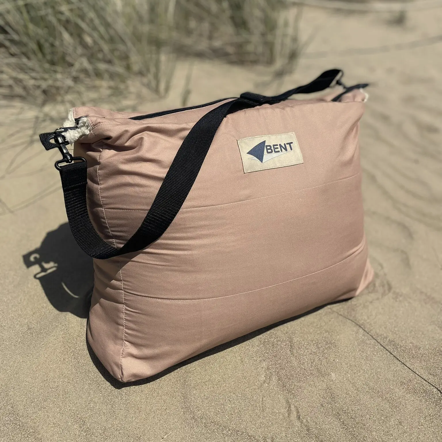 Rosé-beige gepolsterte Schlafsackrolle mit schwarzem Tragegurt liegt im Sand, Logo Bent auf dem Patch sichtbar