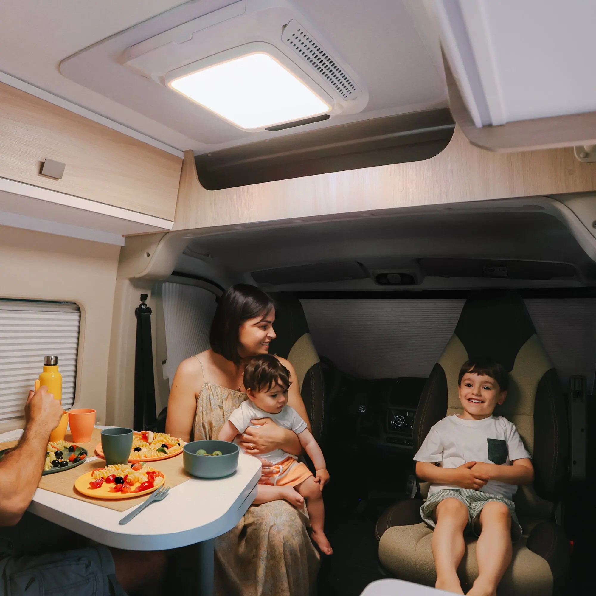 Innenraum eines Wohnmobils mit hellem rechteckigem Deckenlichtpanel und Belüftungsgitter, Esstisch mit Obst, lachende Familie sitzt am Tisch