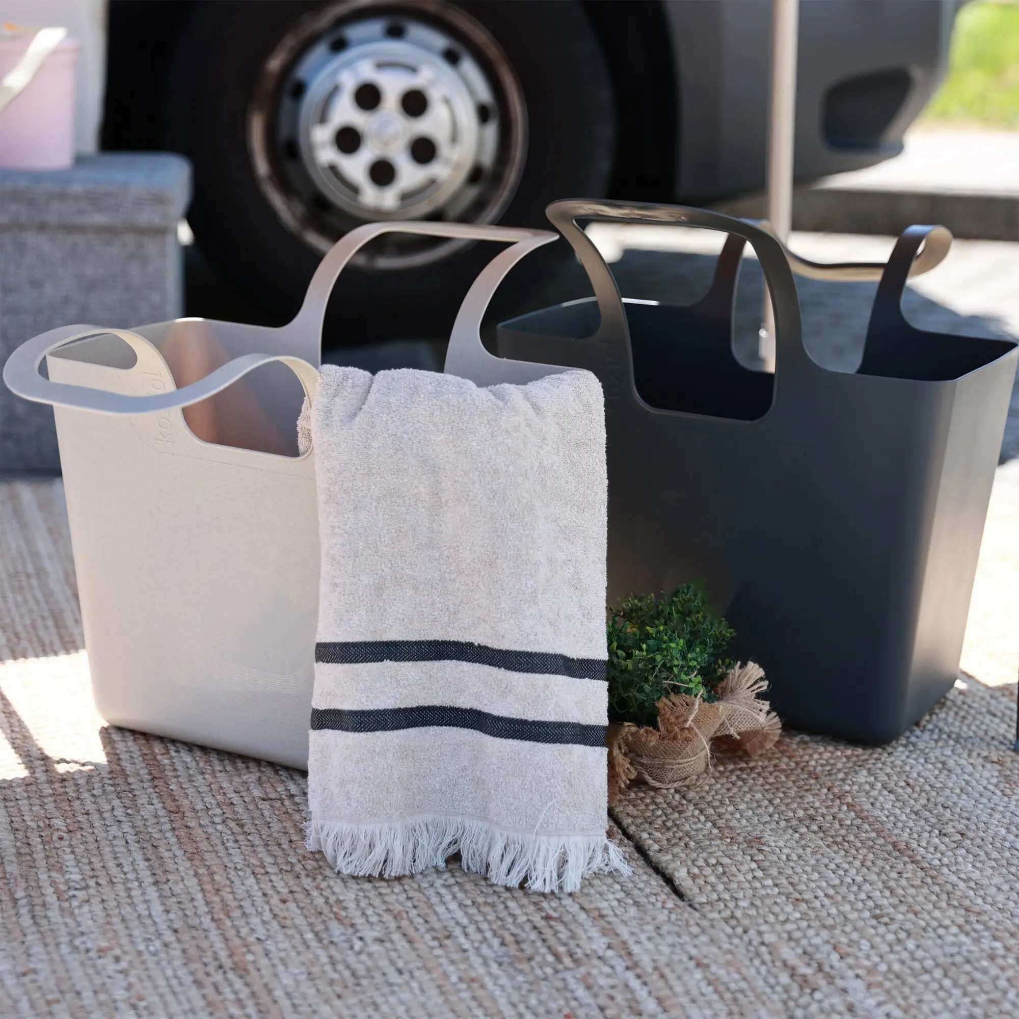 Große graue XL-Tasche mit breiten Griffen, ein weißes Handtuch mit schwarzen Streifen hängt über dem Rand; im Hintergrund ist ein Autoreifen sichtbar