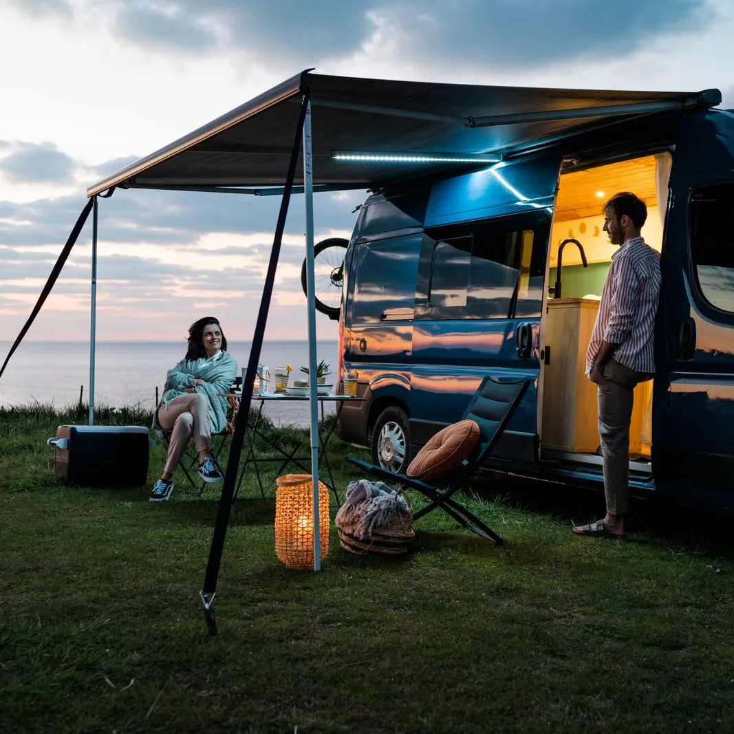 Campingbus mit ausgefahrener Markise und sichtbarer LED-Spannstange Rafter; darunter sitzen zwei Personen bei Abendbeleuchtung