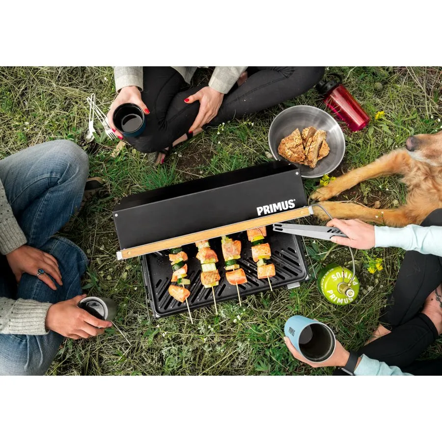 Mobiler schwarzer Primus-Gasgrill Kuchoma mit geöffnetem Deckel, Grillspießen und Grillgut auf einer Wiese in geselliger Runde