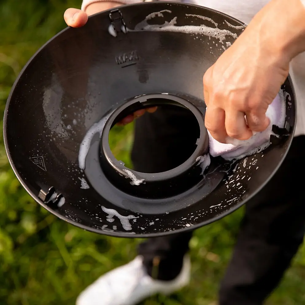 Eine Person wischt ein rundes schwarzes Bauteil des Tischgrill 360 Grill CV mit Seifenlauge und einem Lappen ab, Gras im Hintergrund