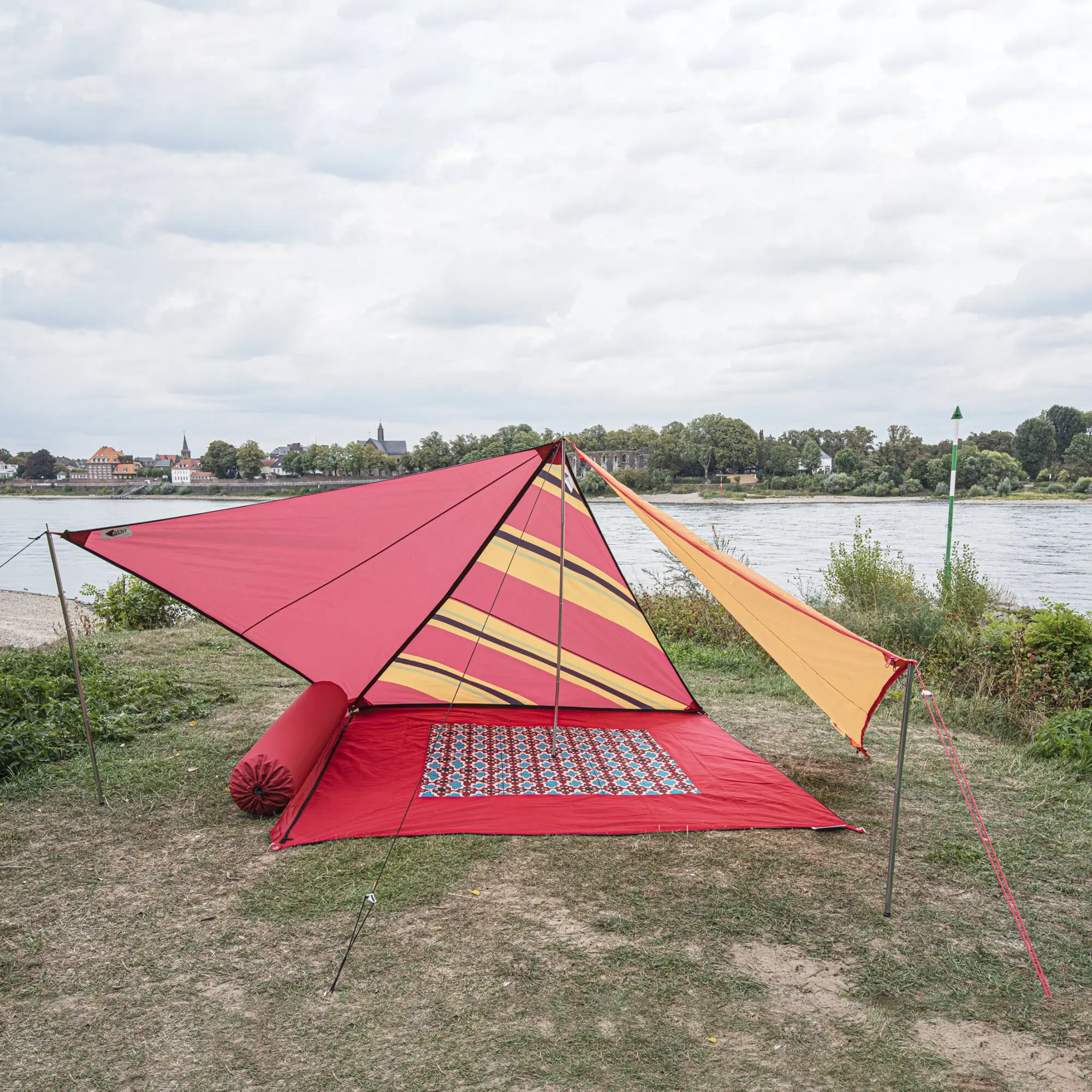 Zip Canvas Sonnensegel mit orange-gelben Streifen über roter Bodenmatte mit geometrischem Teppich; am Ufer befestigt, Hintergrund mit Bäumen