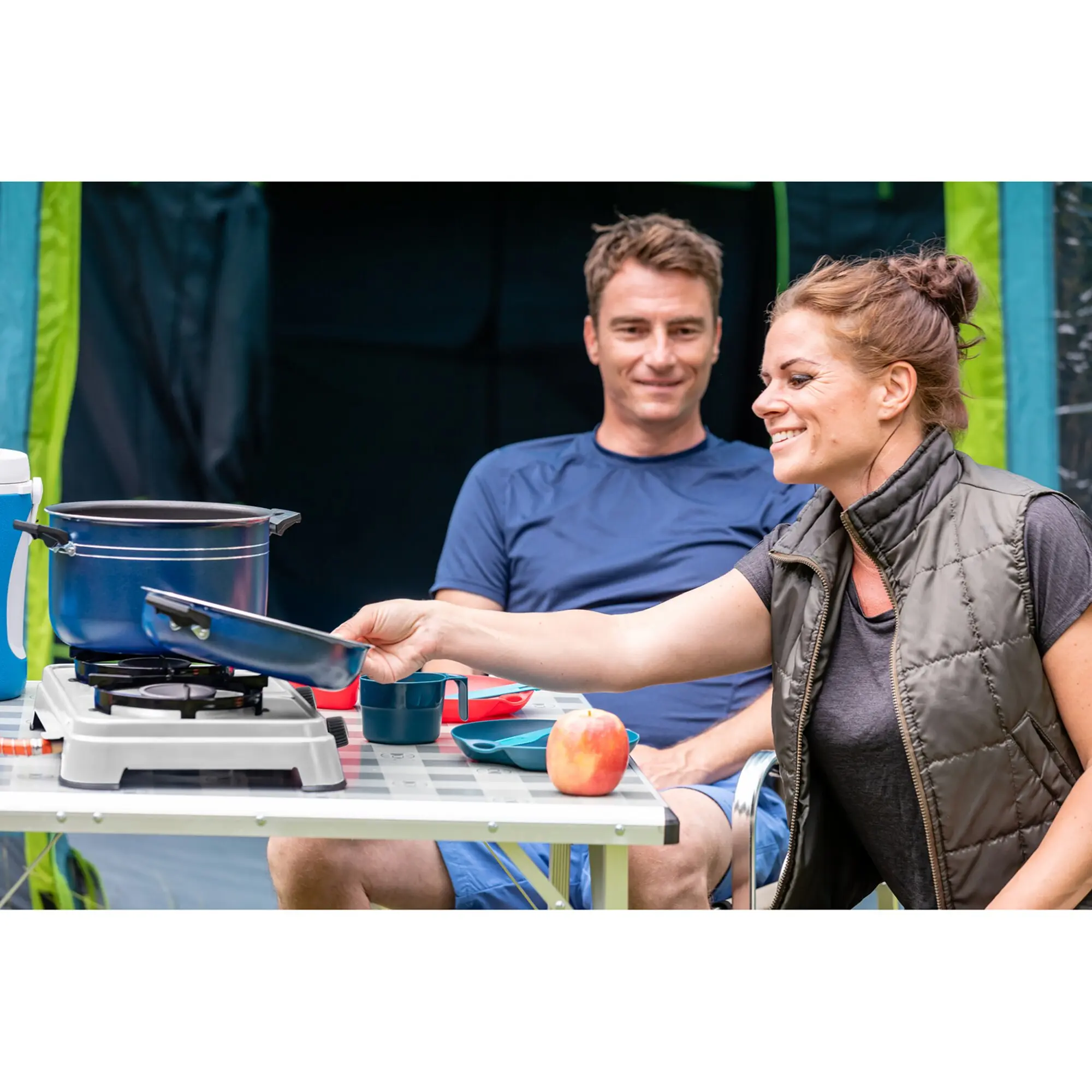 Zwei Personen kochen am Campingtisch mit dem Camping Cook CV Kocher, blauem Topf, daneben Apfel und blaues Geschirr, grünes Zelt