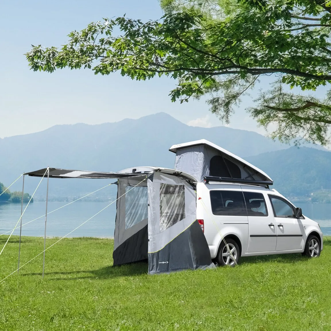 Graues Heckzelt Pilote Caddy 5 an einem weißen Caddy mit Markise auf einer Wiese am See vor Bergkulisse