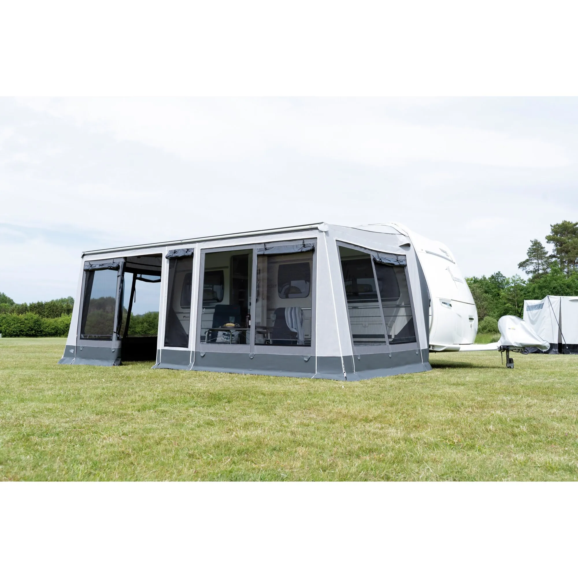 Graues Markisenzelt Rolli Plus Lounge an einem weißen Wohnwagen auf einer Campingwiese, mit großen transparenten Fensterflächen und geöffnetem Eingang